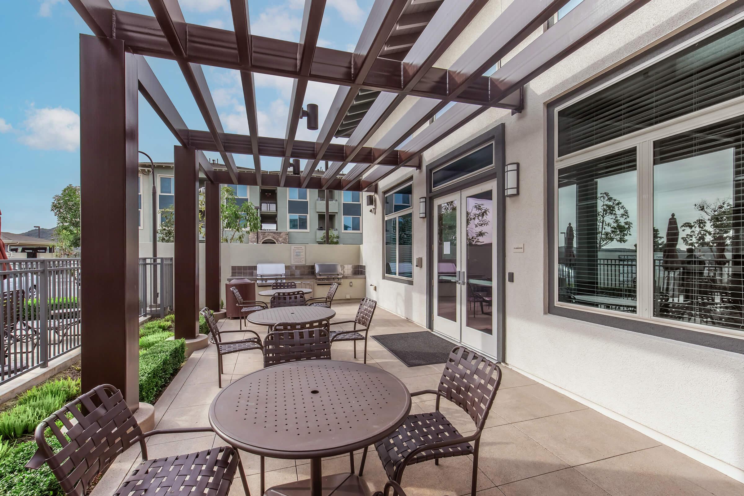 An outdoor patio area featuring multiple round tables with chairs, shaded by a pergola. The space is surrounded by greenery, and there are windows showing a modern building in the background. Bright blue sky with a few clouds is visible.