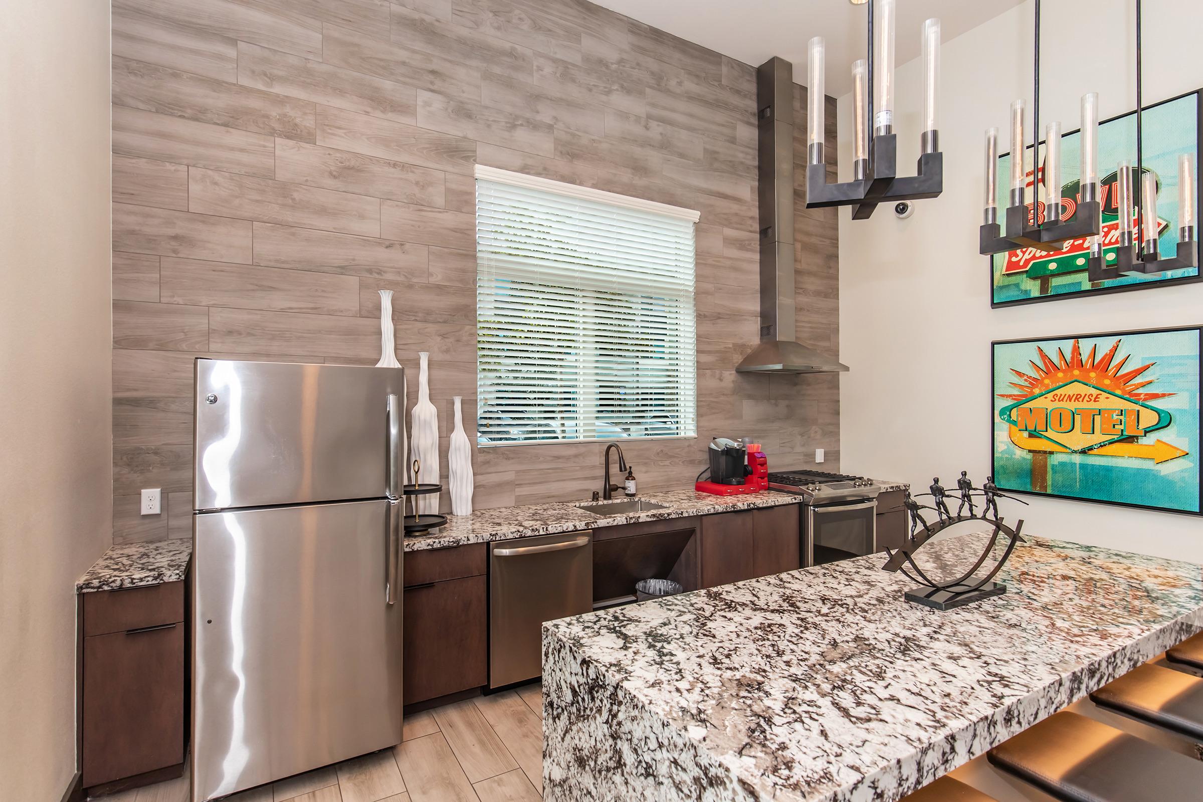 Modern kitchen featuring stainless steel appliances, including a refrigerator and a dishwasher, complemented by dark cabinetry and a grey marble countertop. Natural light streams in through a large window, illuminating the space, which includes decorative elements like vases and wall art.