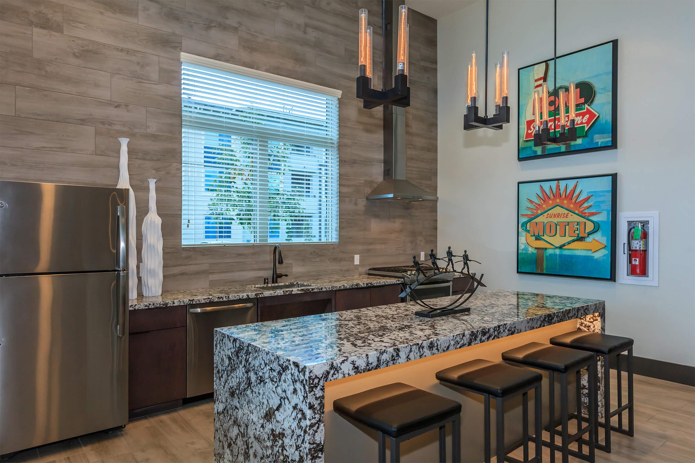 Modern kitchen with a granite countertop and dark cabinets. It features stylish bar stools, a stainless steel refrigerator, and a hanging light fixture. Decorative wall art includes vibrant signs. Large windows allow natural light to illuminate the space.