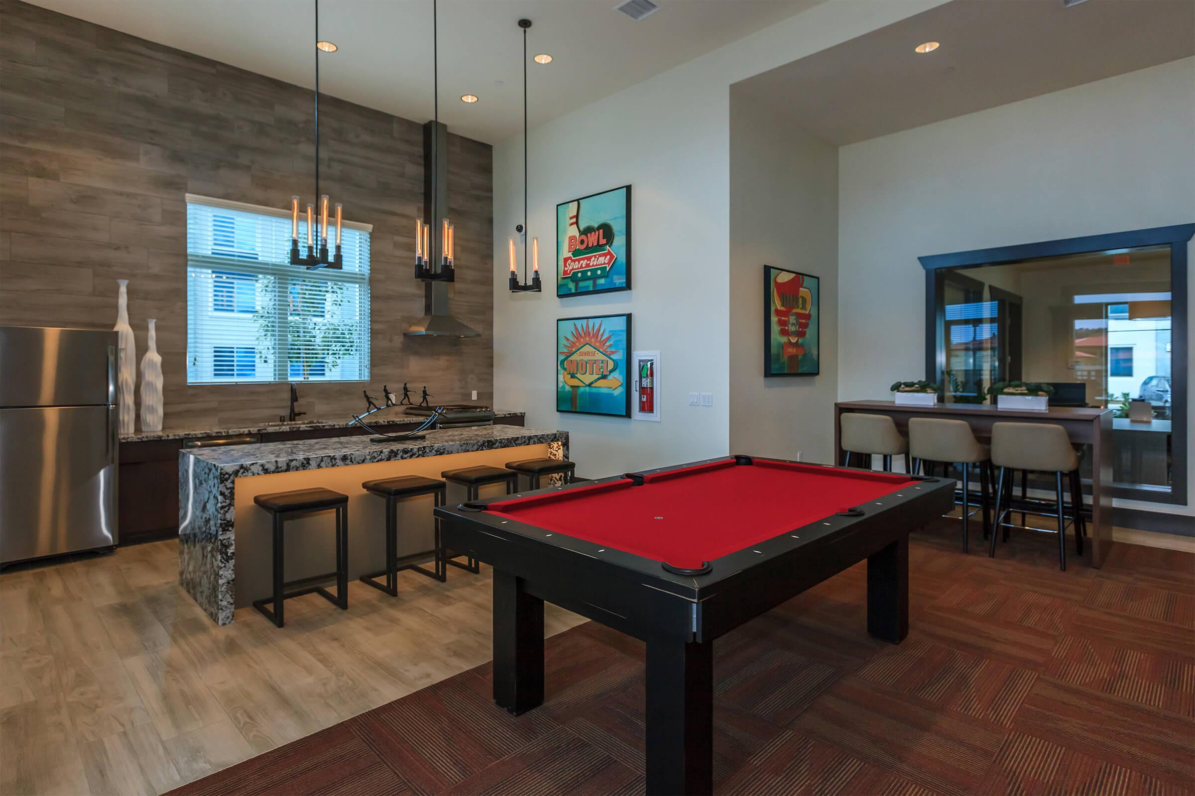 A modern recreational room featuring a red pool table, a bar area with high stools, and decorative wall art. The space has large windows, a stylish kitchen with granite countertops, and pendant lighting, creating a welcoming atmosphere for social gatherings.