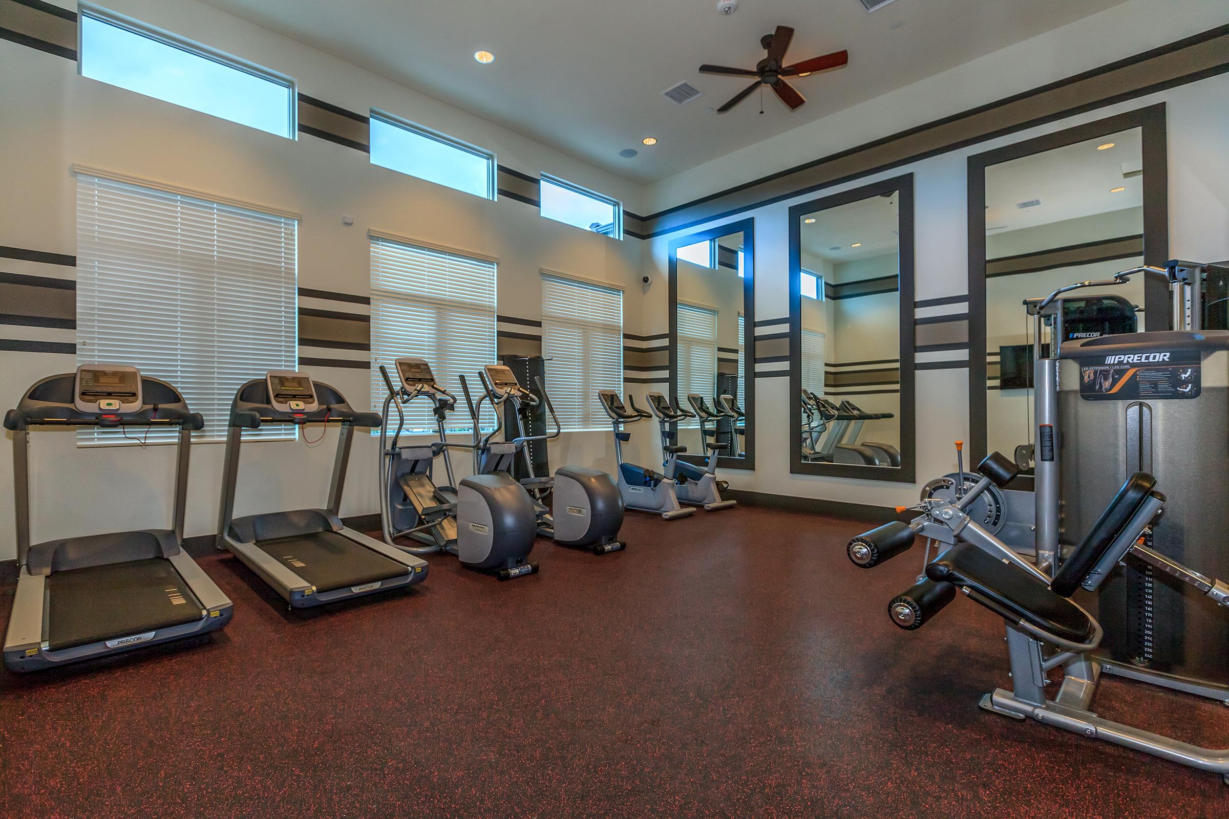 A spacious gym with multiple treadmills, exercise bikes, and a strength training machine. Large windows provide natural light, while mirrors enhance the sense of space. The floor is covered with dark rubber flooring, and ceiling fans are visible, creating a comfortable workout environment.