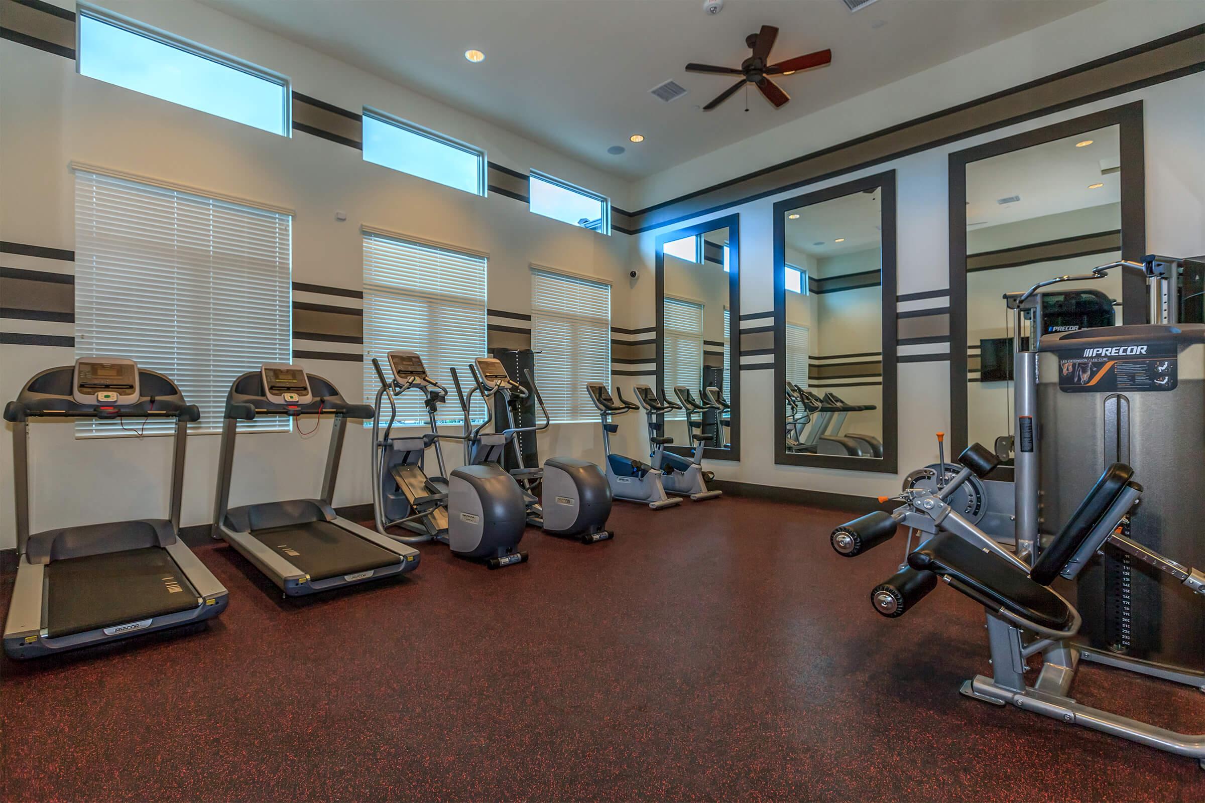 A modern fitness room featuring two treadmills, stationary bikes, and a multi-gym machine. The space has large windows for natural light, mirrors on the walls, and a dark rubber flooring. The clean and well-lit environment is designed for a comfortable workout experience.
