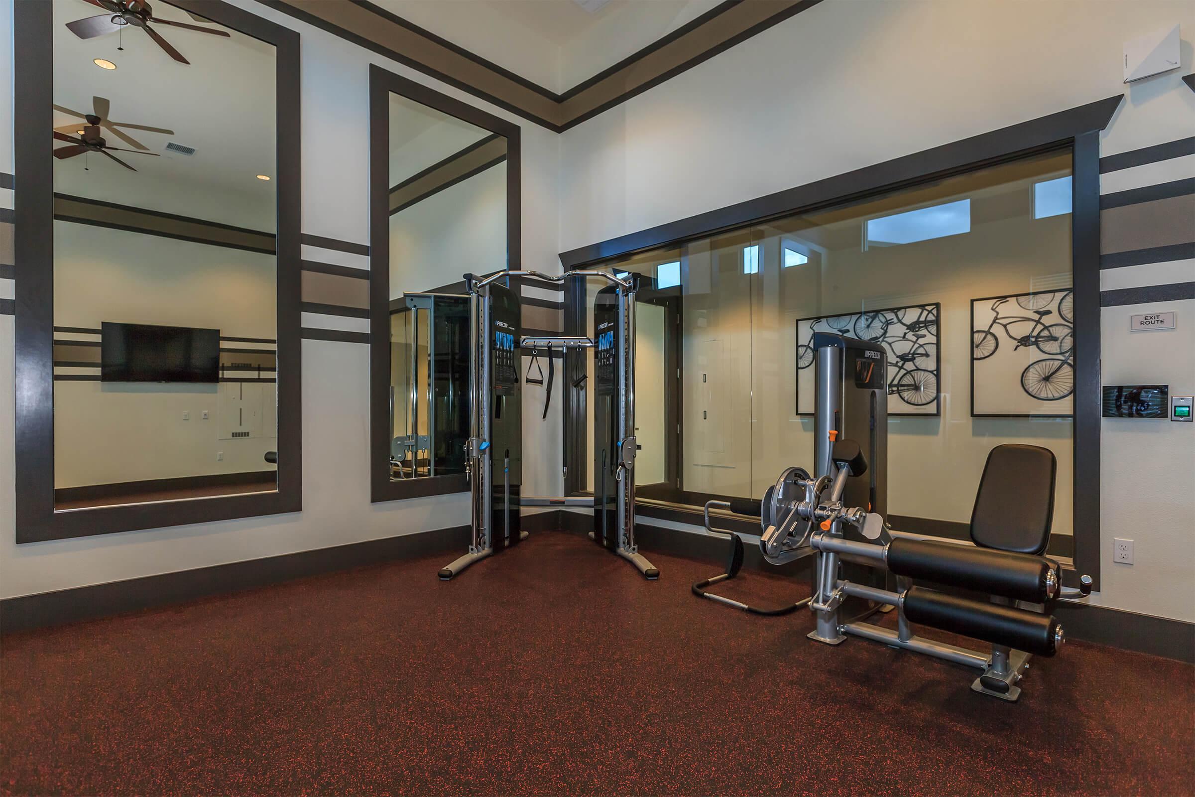 A modern gym space featuring a multi-station workout machine, a bench press, and exercise equipment. Large mirrors reflect the room, which has a sleek design with dark accents and an inviting atmosphere. Wall art includes framed images of bicycles, enhancing the fitness theme.