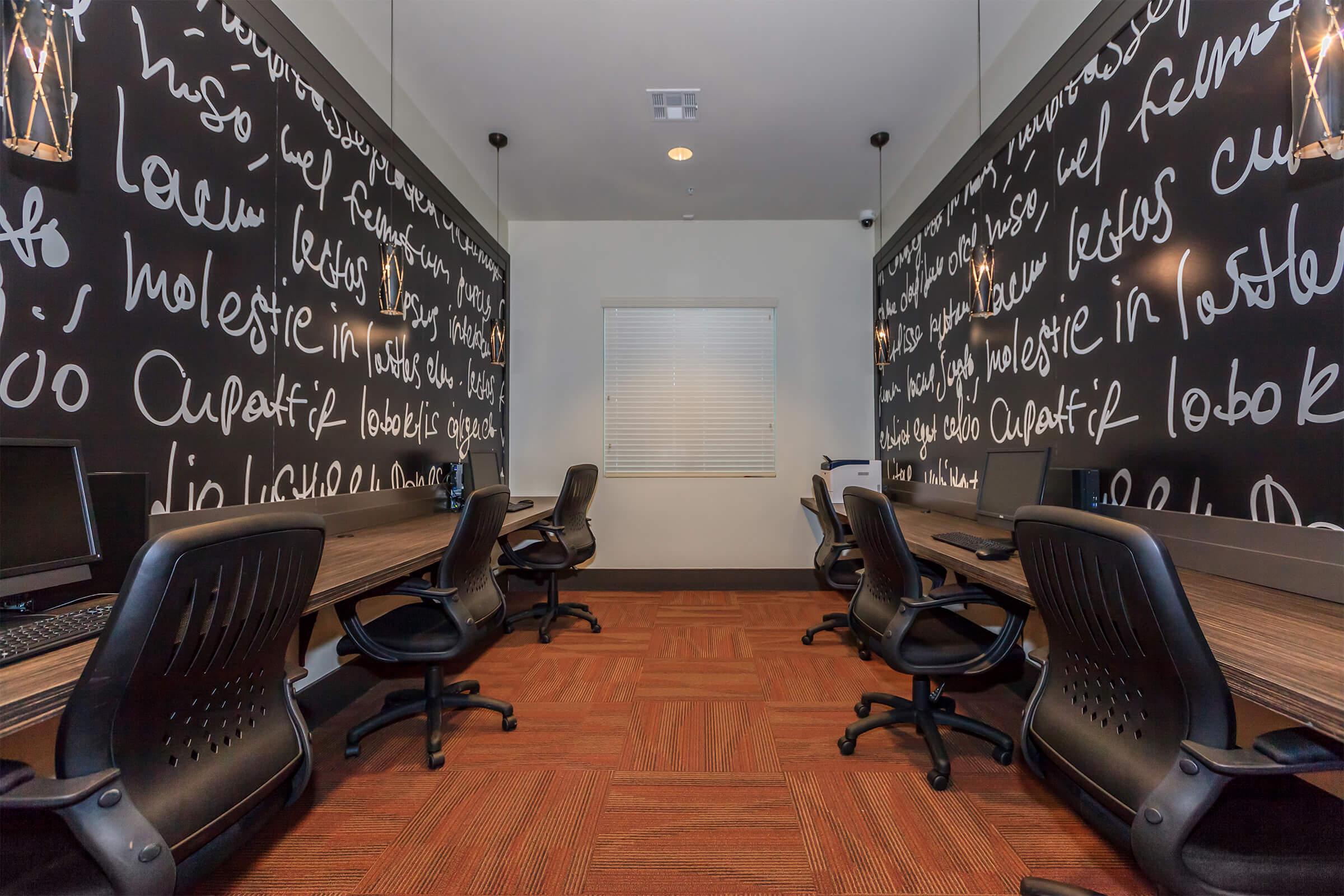 A study room with two long tables and black chairs, featuring walls decorated with white handwritten text. There are several computer desks arranged neatly, and a window with closed blinds letting in soft light. Modern pendant lights hang from the ceiling, creating a focused and collaborative environment.