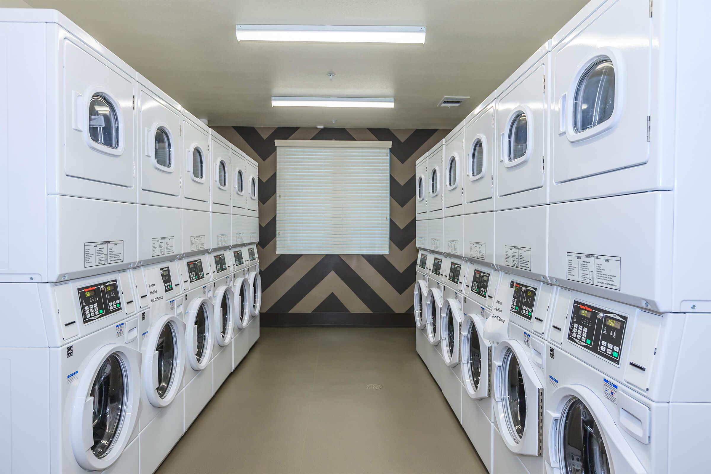 A clean and modern laundry room featuring rows of white washers and dryers, with a window covered by a light-colored blind. The walls have a stylish chevron pattern, and overhead lighting provides bright illumination for the space.