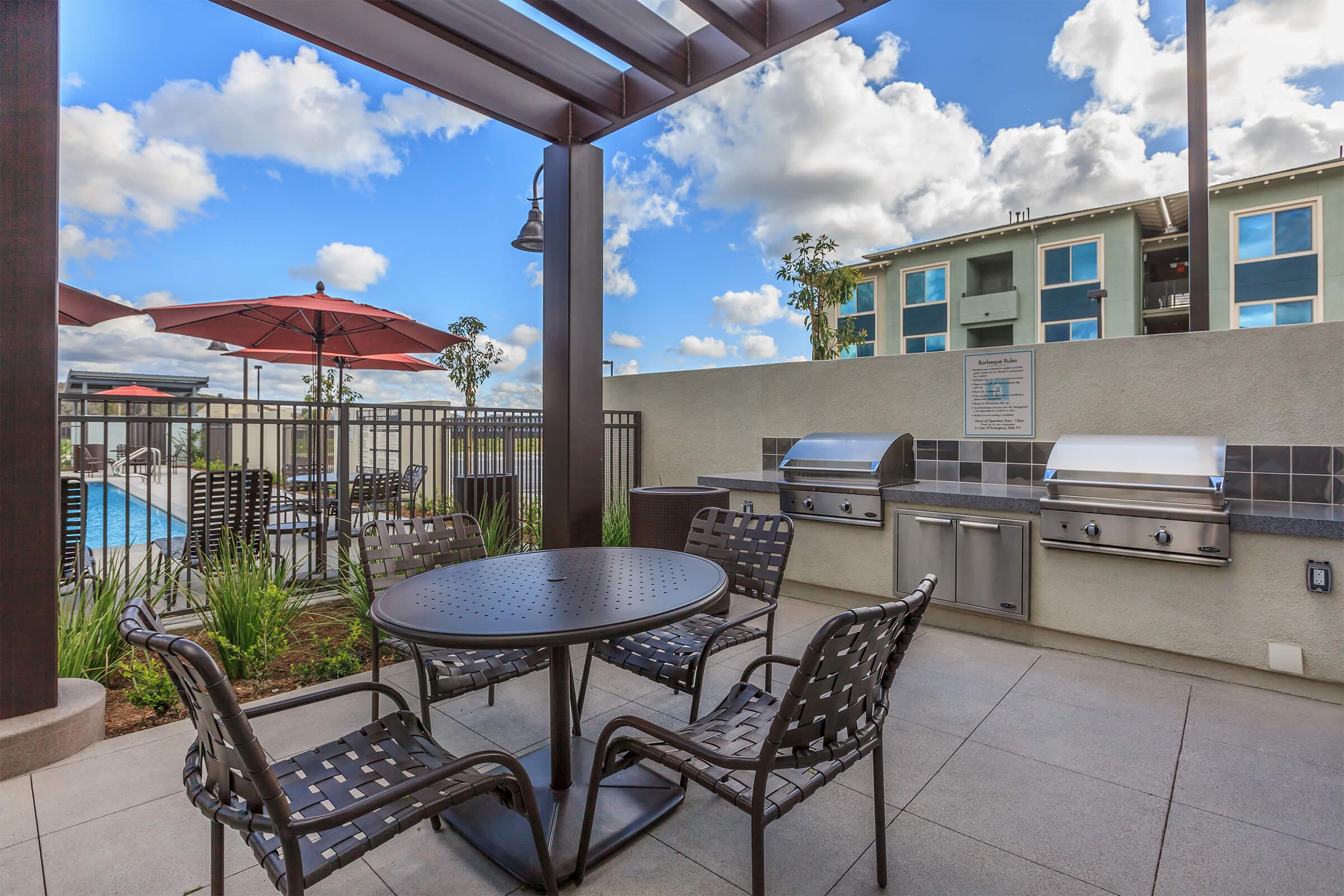 Outdoor dining area featuring a round table with four chairs under a shaded pergola. Two BBQ grills are positioned against a wall beside the table. In the background, there are umbrellas over additional seating and a glimpse of a pool area. The sky is partly cloudy.
