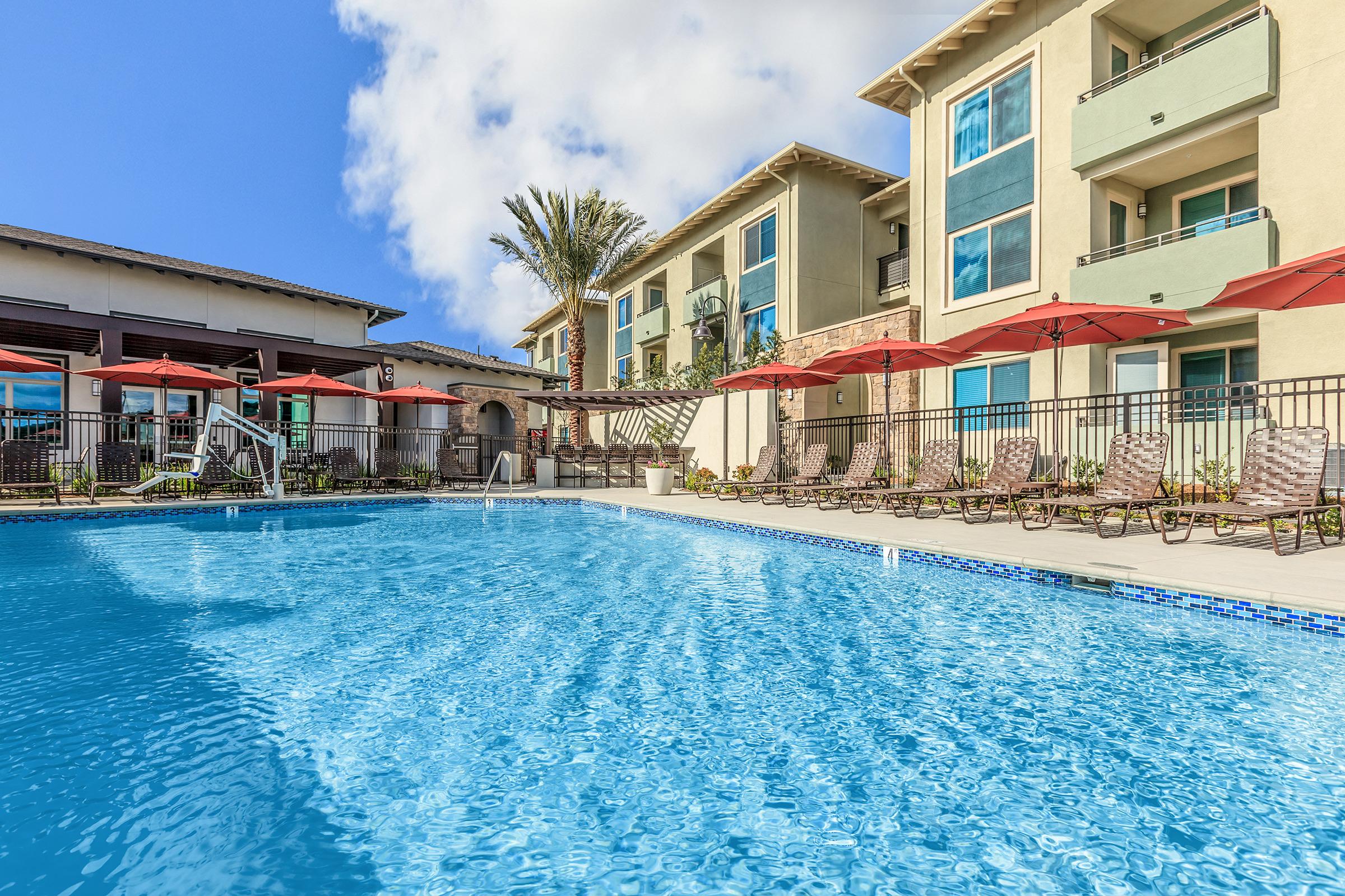 A crystal-clear swimming pool surrounded by lounge chairs and red umbrellas, with modern apartment buildings in the background. The scene features palm trees and a bright blue sky, creating a relaxing and inviting atmosphere.