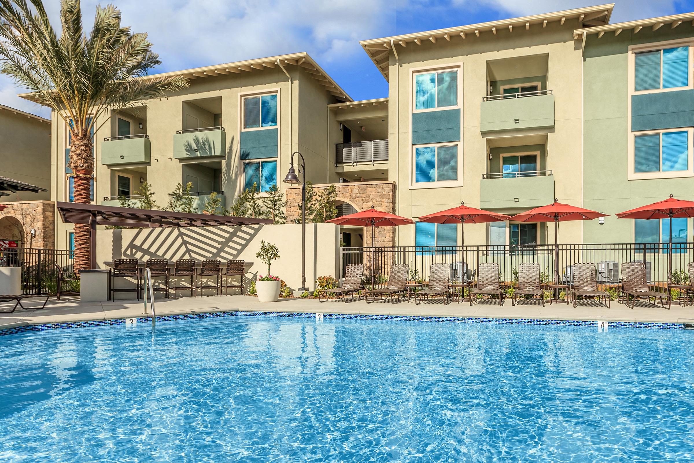 A swimming pool surrounded by lounge chairs and umbrellas, with modern apartment buildings in the background. The scene features palm trees and a clear blue sky, creating a relaxing atmosphere.