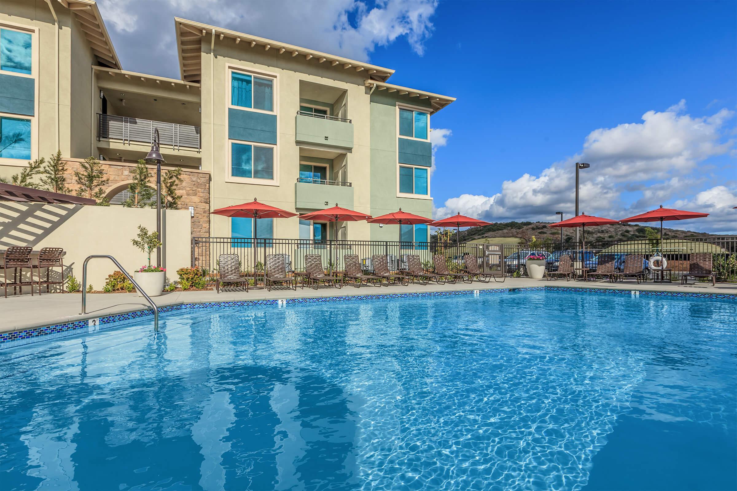 A clear blue swimming pool surrounded by lounge chairs and red umbrellas, with a modern apartment building in the background. The sky is bright with a few clouds, creating a serene ambiance, ideal for relaxation and leisure.