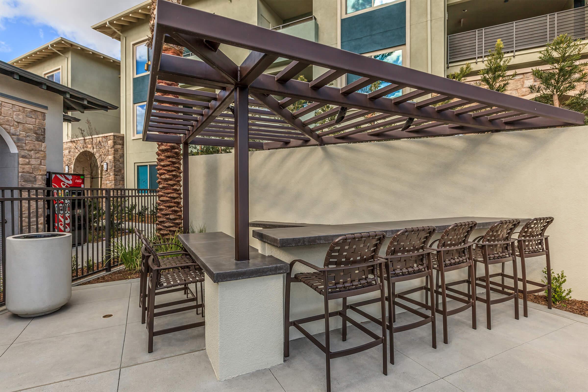 A shaded outdoor seating area featuring a long, dark metal bar with high chairs, set against a modern building facade. The space is surrounded by landscaping and a fence, creating a cozy atmosphere for relaxation or social gatherings.
