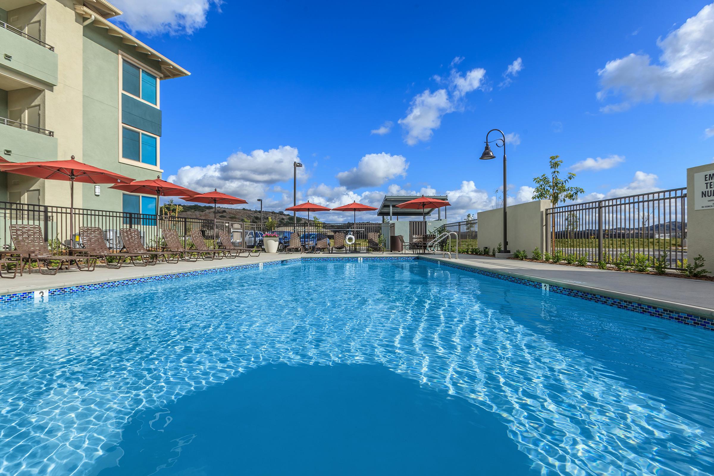 A bright blue swimming pool surrounded by lounge chairs and red umbrellas. In the background, a modern building with large windows is visible under a clear blue sky with fluffy white clouds. A landscaped area with greenery is also present near the pool.