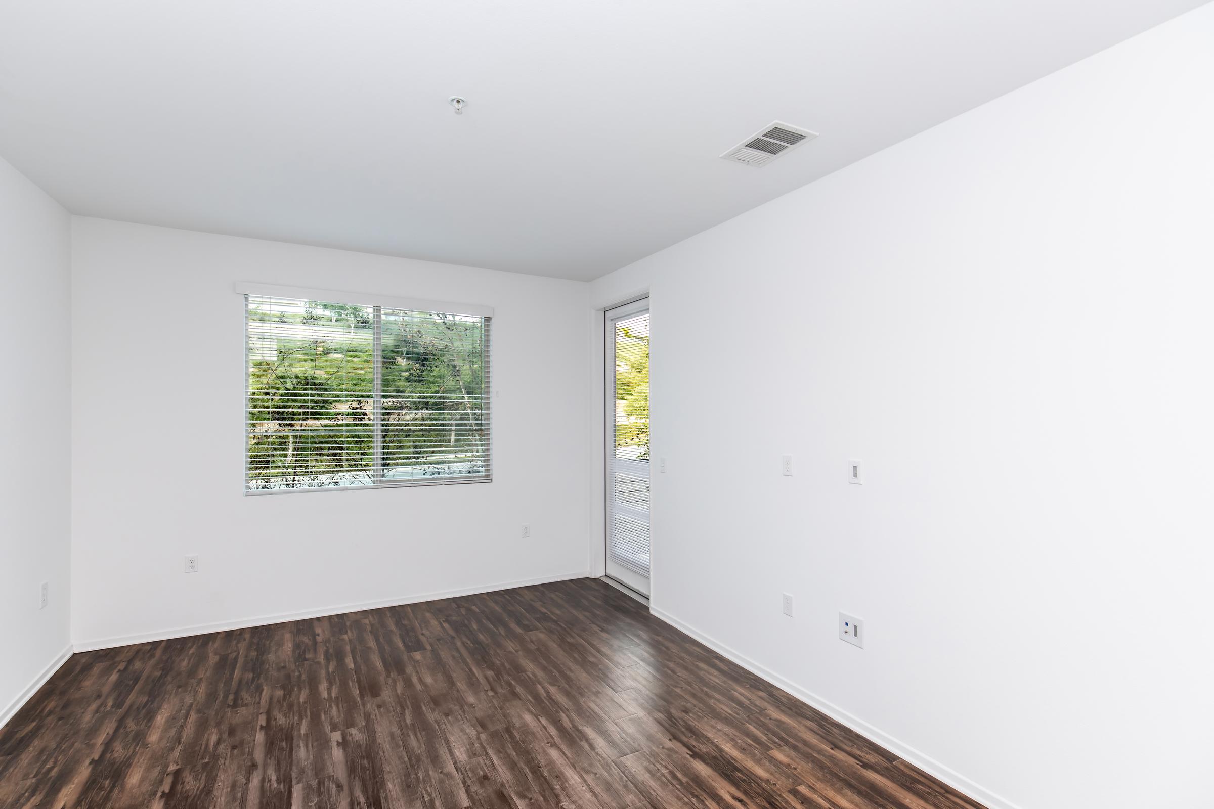 A bright, empty room with white walls and a large window featuring blinds. The floor is dark, resembling wood. There’s a sliding glass door to the right, leading outside. The space is minimalist and uncluttered, with plenty of natural light.