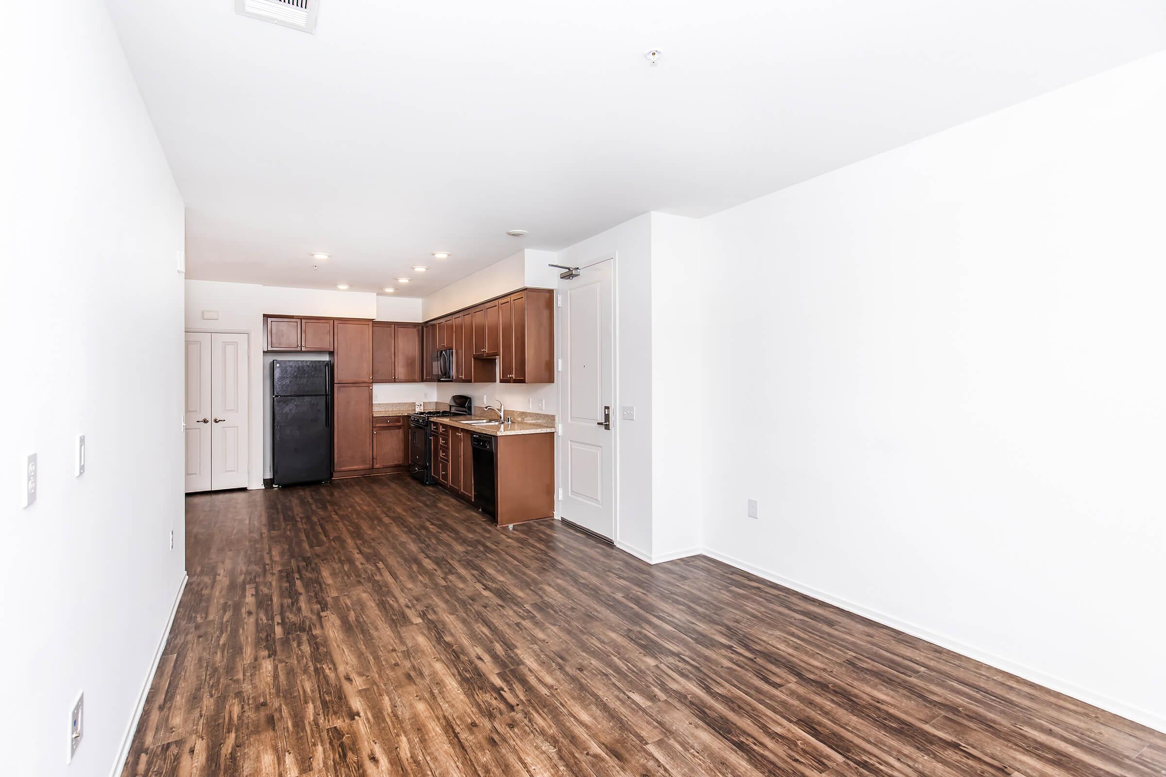 Modern kitchen and living space with dark wood cabinets, granite countertops, and stainless steel appliances. The room features hardwood flooring and white walls, providing a clean, open look with natural light. The kitchen is partially visible with a black refrigerator and oven.