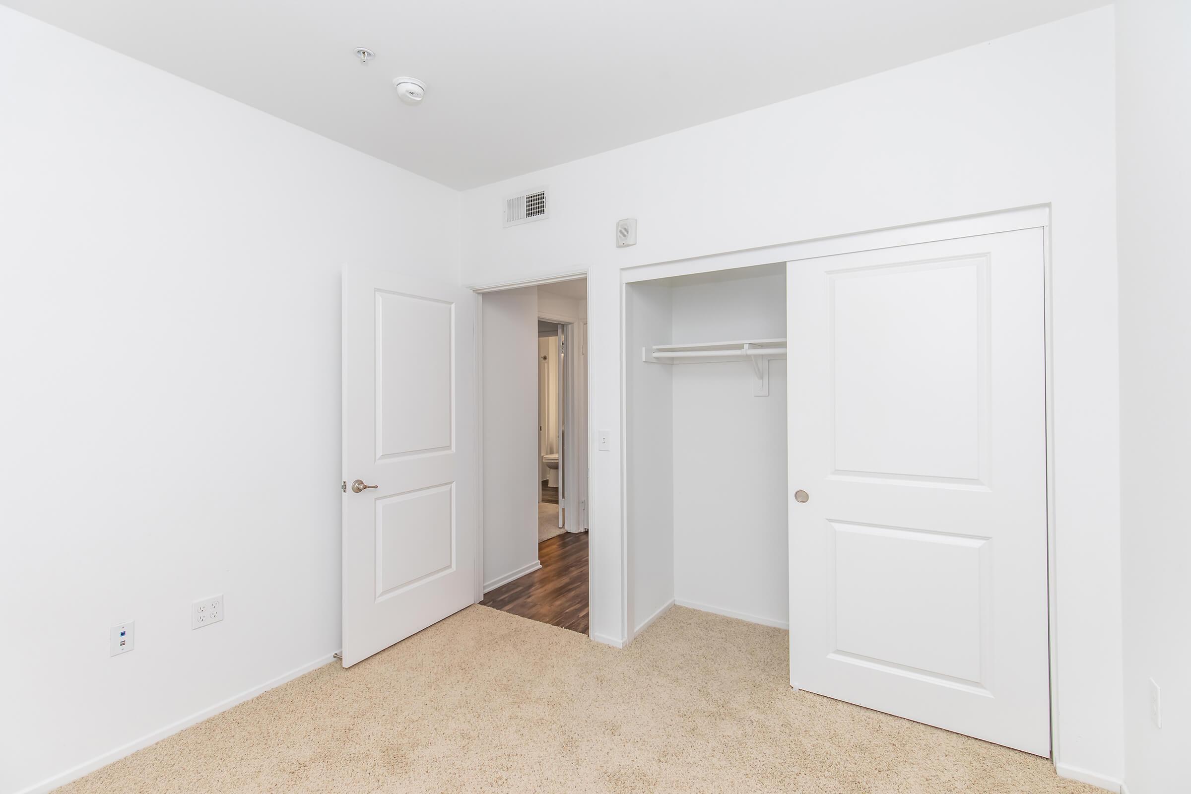A bright, empty room with light beige carpet. There are two white doors, one leading to a closet and the other to an adjoining space. The walls are painted white, and there is a ceiling vent. The overall atmosphere is clean and minimalistic.