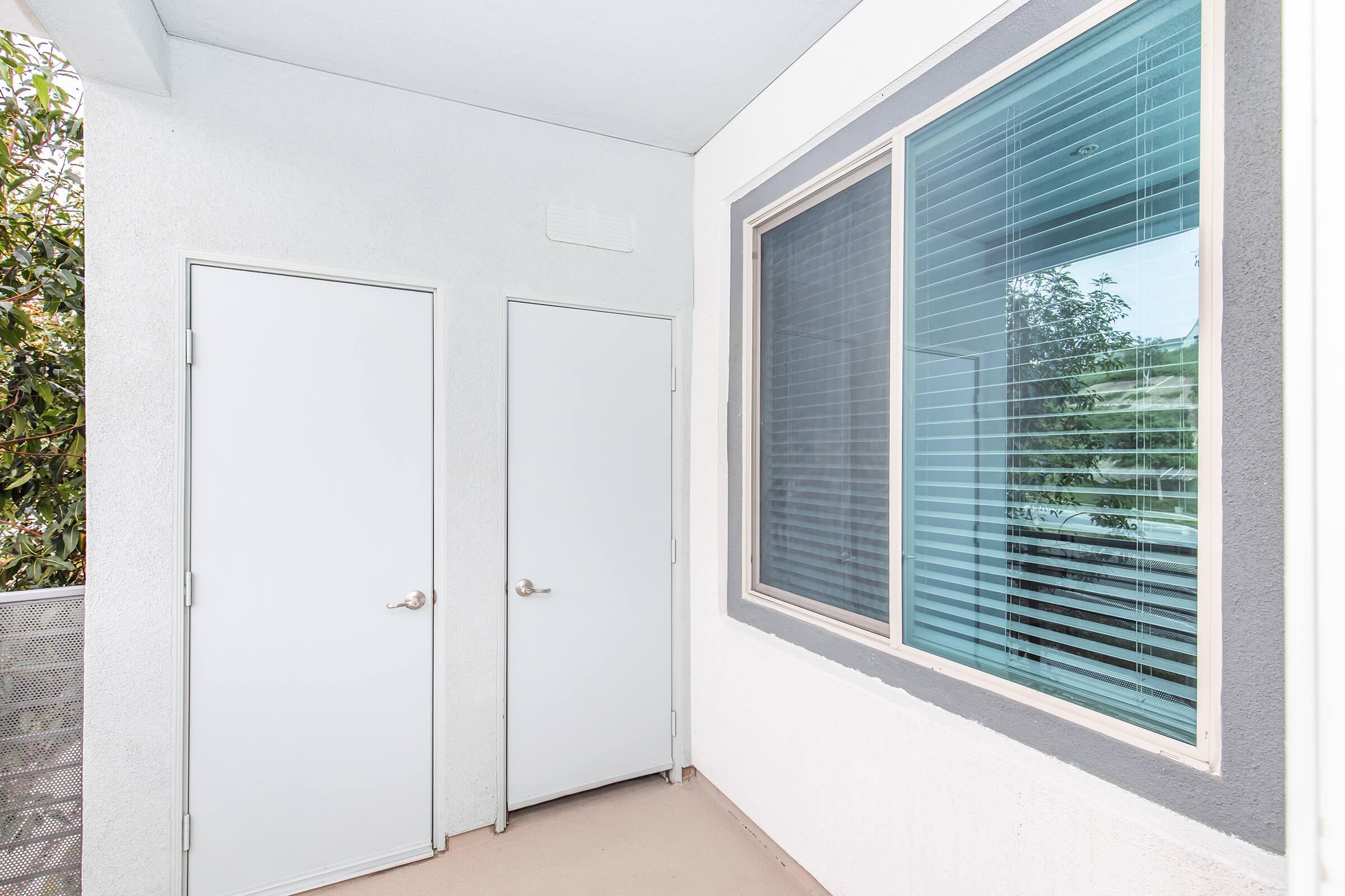 Two white doors are set in a light-colored wall, next to a large window with horizontal blinds. The area appears well-lit and clean, with greenery partially visible outside the window. The floor is neutral in color, and the overall space has a modern and minimalistic design.