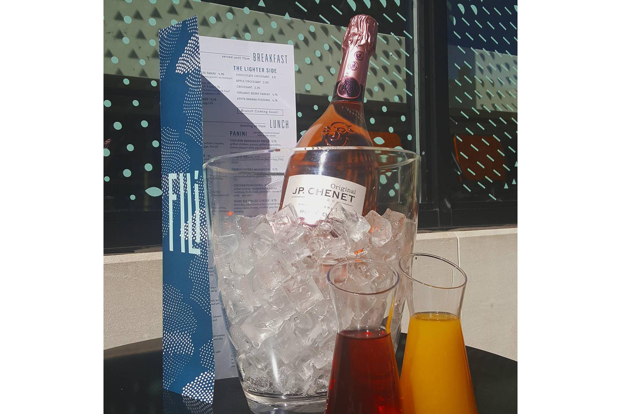 A bottle of rosé champagne in a bucket filled with ice, accompanied by two glasses of colorful drinks—one red and one orange. A menu is visible in the background, featuring options for breakfast and lunch. The setting has a modern vibe, with a patterned backdrop.