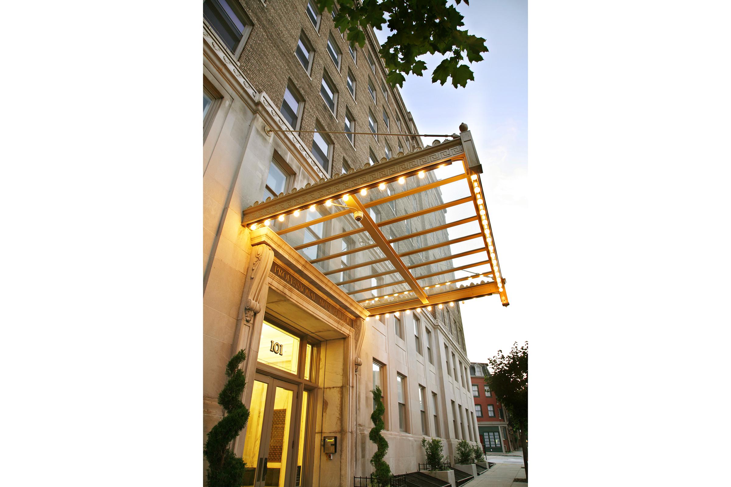A building entrance featuring a decorative awning with warm lighting. The awning is supported by a metallic frame and extends outward from the entrance. The building has a brick facade and several windows, with greenery partially framing the entrance. The scene is set during twilight, creating a warm ambiance.