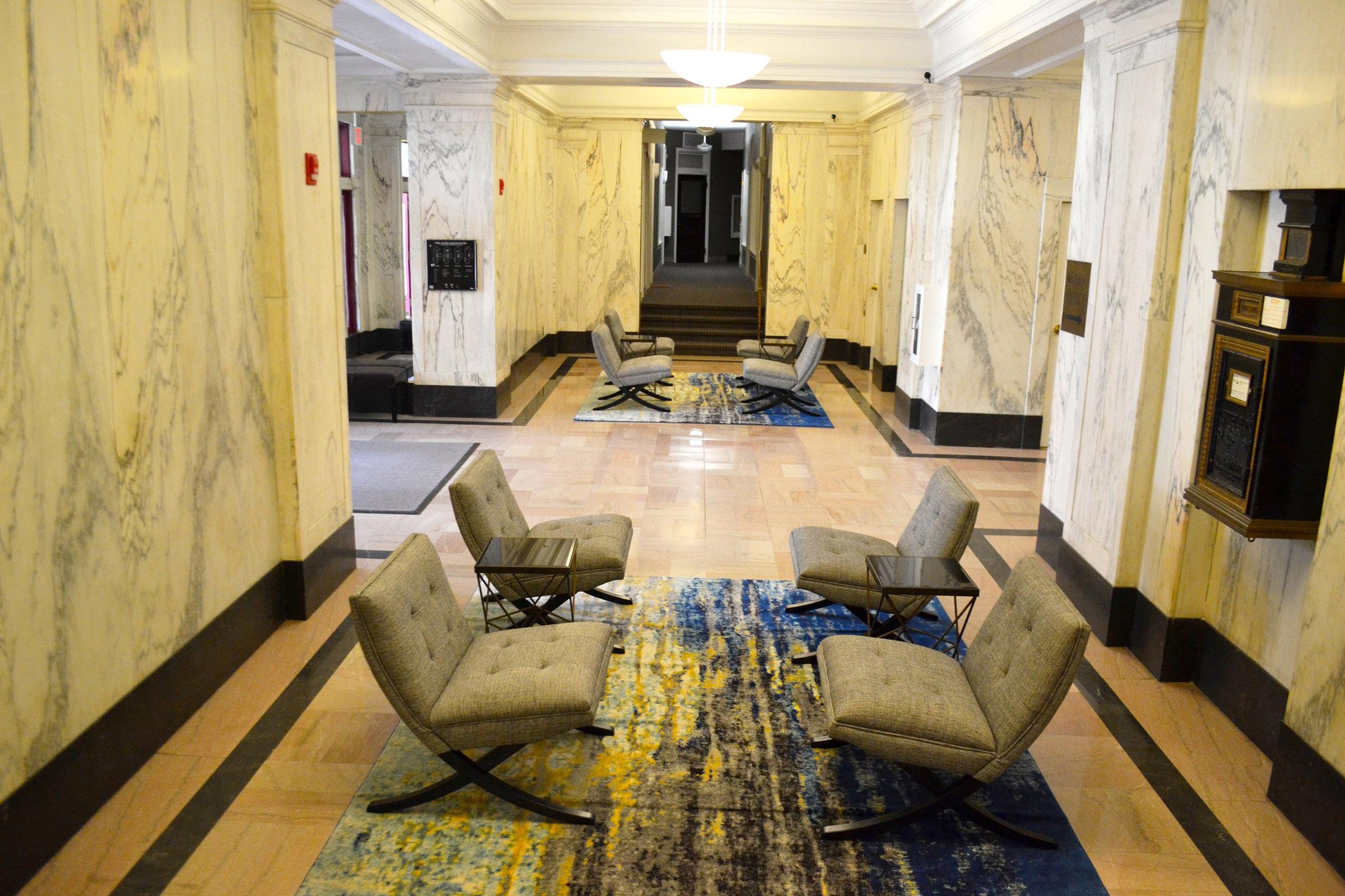 A spacious indoor corridor featuring marble walls and a polished floor. Two sets of modern chairs with small tables are placed on a colorful area rug. The hallway is illuminated by overhead lighting, creating a bright and inviting atmosphere.