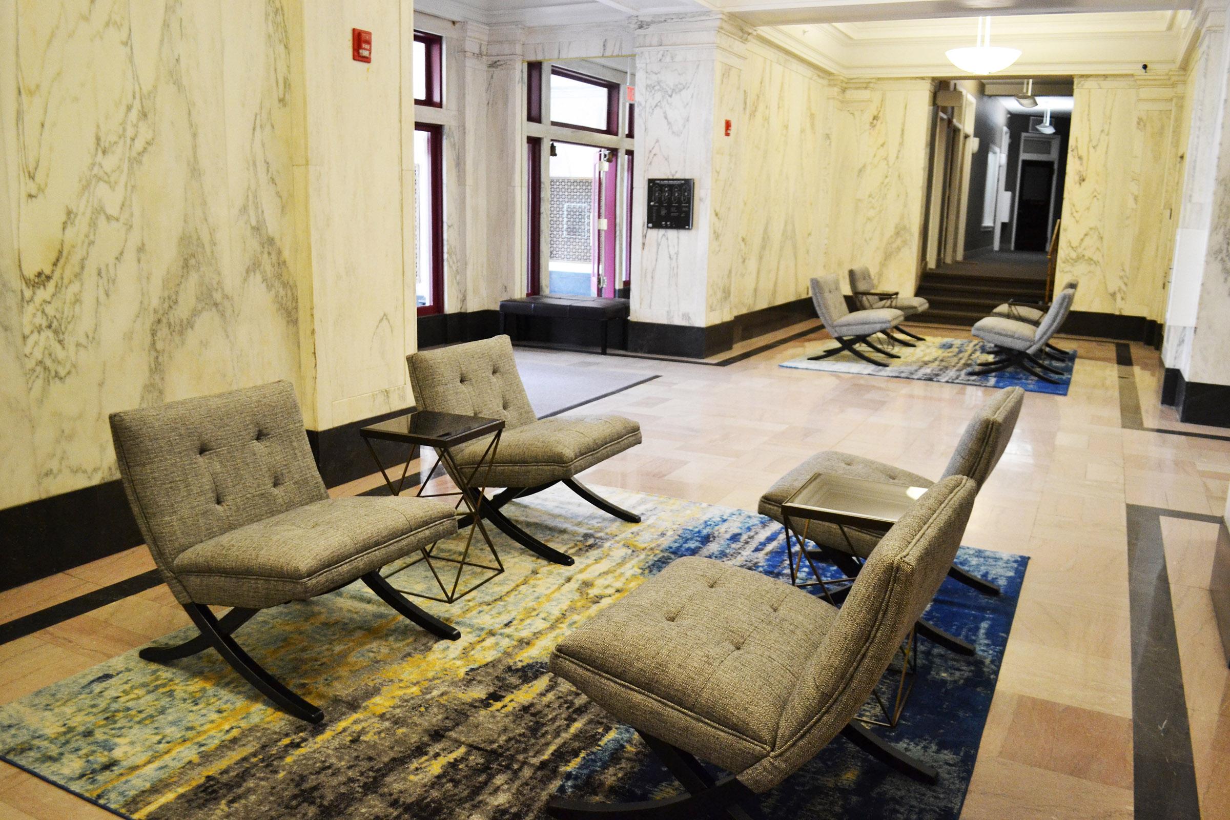 A spacious interior with elegant marble walls and polished floors. Several modern gray lounge chairs are arranged on decorative area rugs, creating a comfortable seating area. Natural light filters in through large windows, enhancing the inviting ambiance of the space.