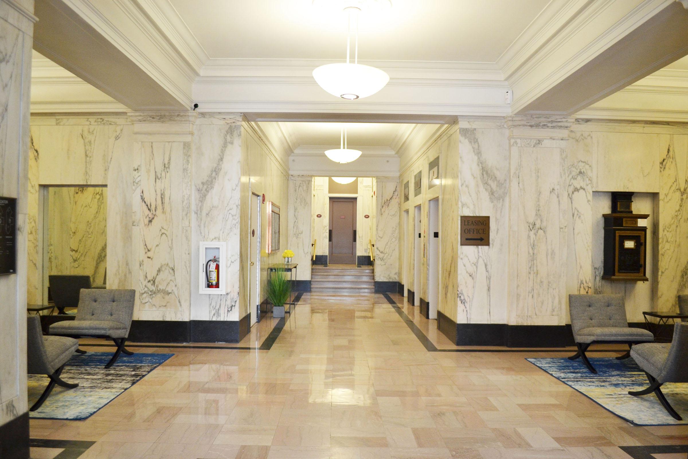 Interior of a marble-finished hallway with two elevators and seating areas on either side. The floor is tiled, featuring a mix of cream and dark colors. Bright overhead lighting illuminates the space, and decorative rugs add color. A potted plant and wall-mounted art are visible, suggesting a welcoming atmosphere.