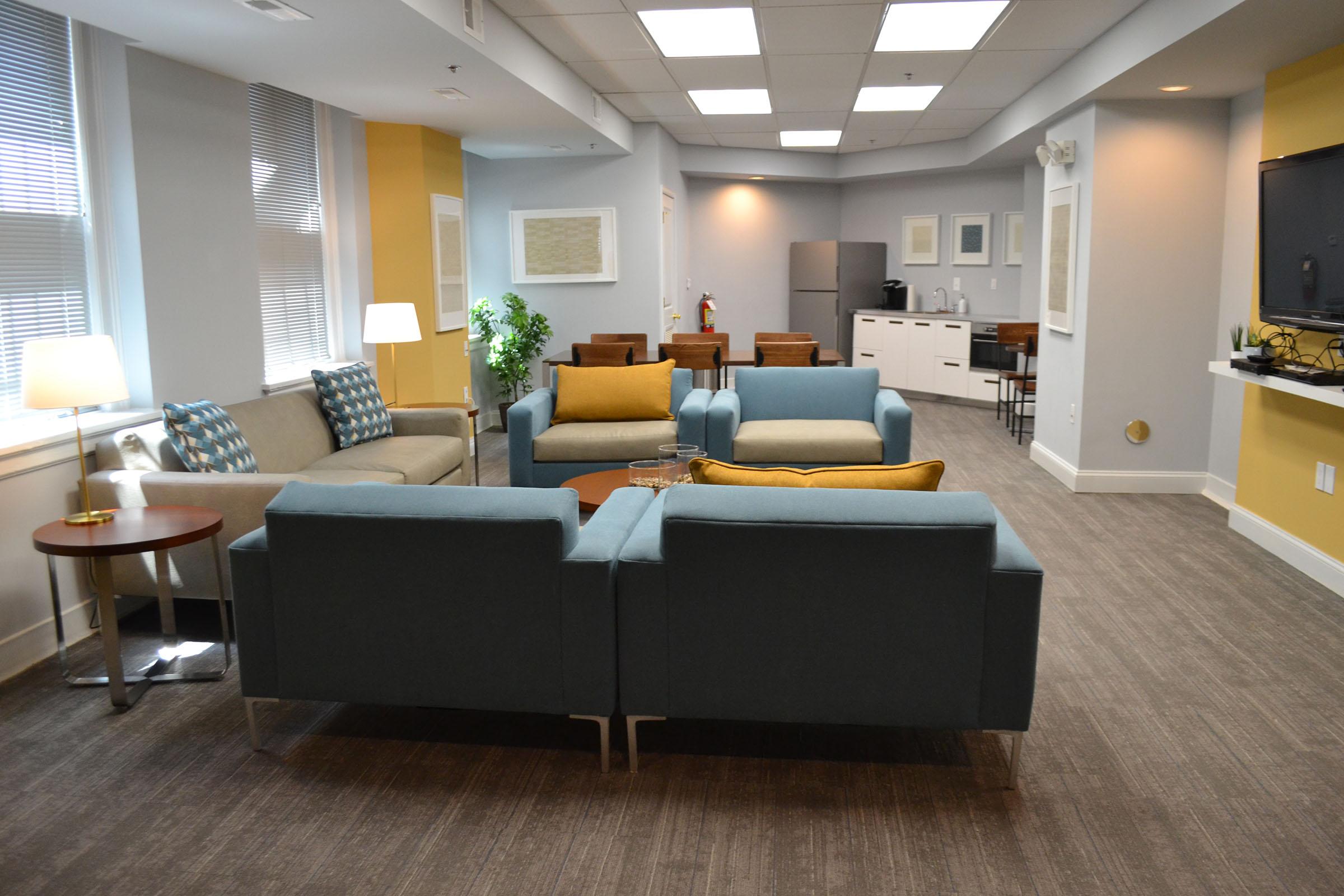 A communal living area featuring modern furniture, including sofas and armchairs in blue and yellow colors. There are lamps for illumination and a kitchenette visible in the background, with a dining area. The space has a cozy, inviting atmosphere with large windows allowing natural light.