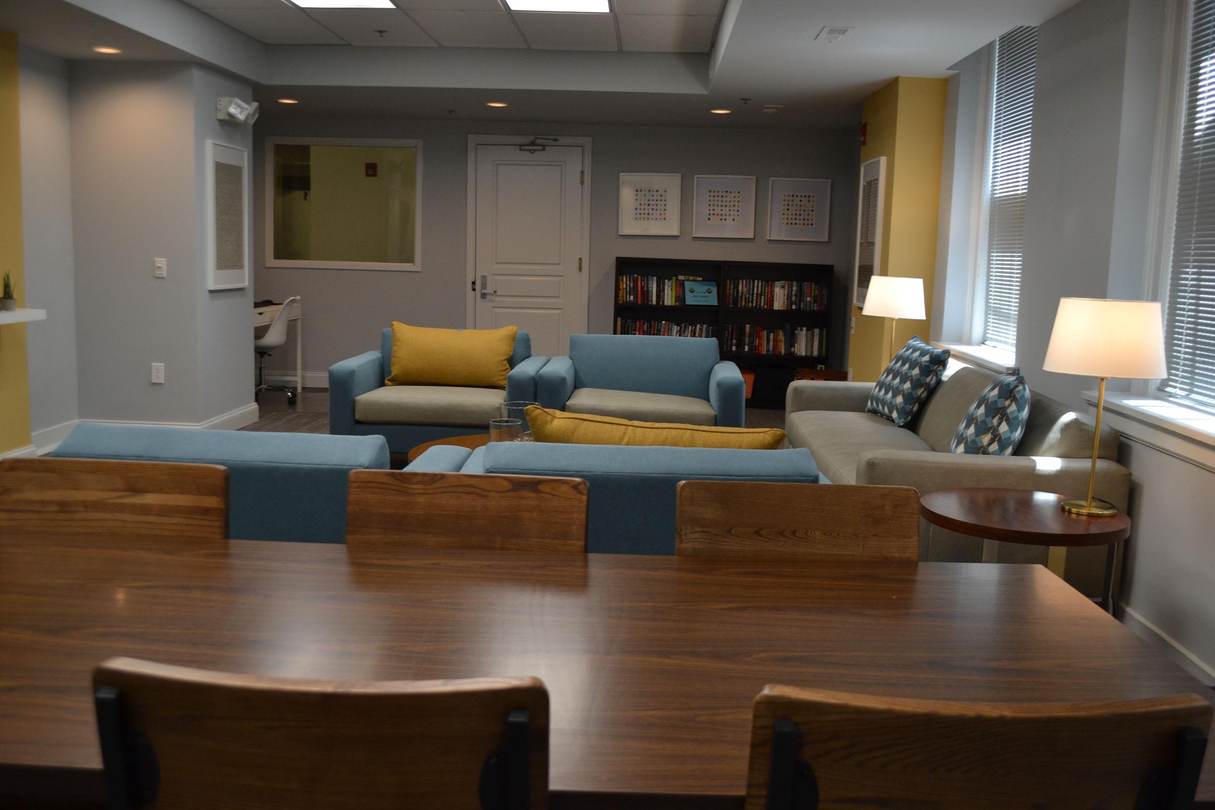 A cozy lounge area featuring blue and gray sofas, a coffee table, and a wooden dining table with chairs. A lamp and bookshelves filled with books add to the inviting atmosphere, along with large windows allowing natural light into the space.