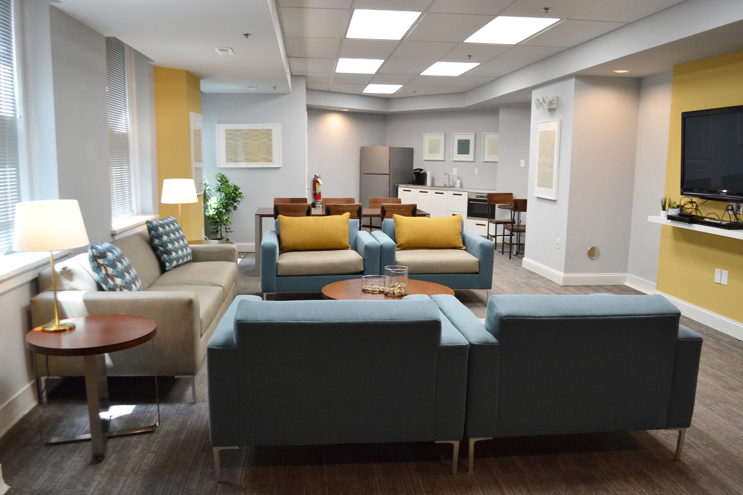 A modern lounge area featuring two sofas, a coffee table, and decorative cushions. A floor lamp provides warm lighting. In the background, a kitchenette with a refrigerator and dining area is visible, with beige and yellow walls creating a cozy atmosphere.