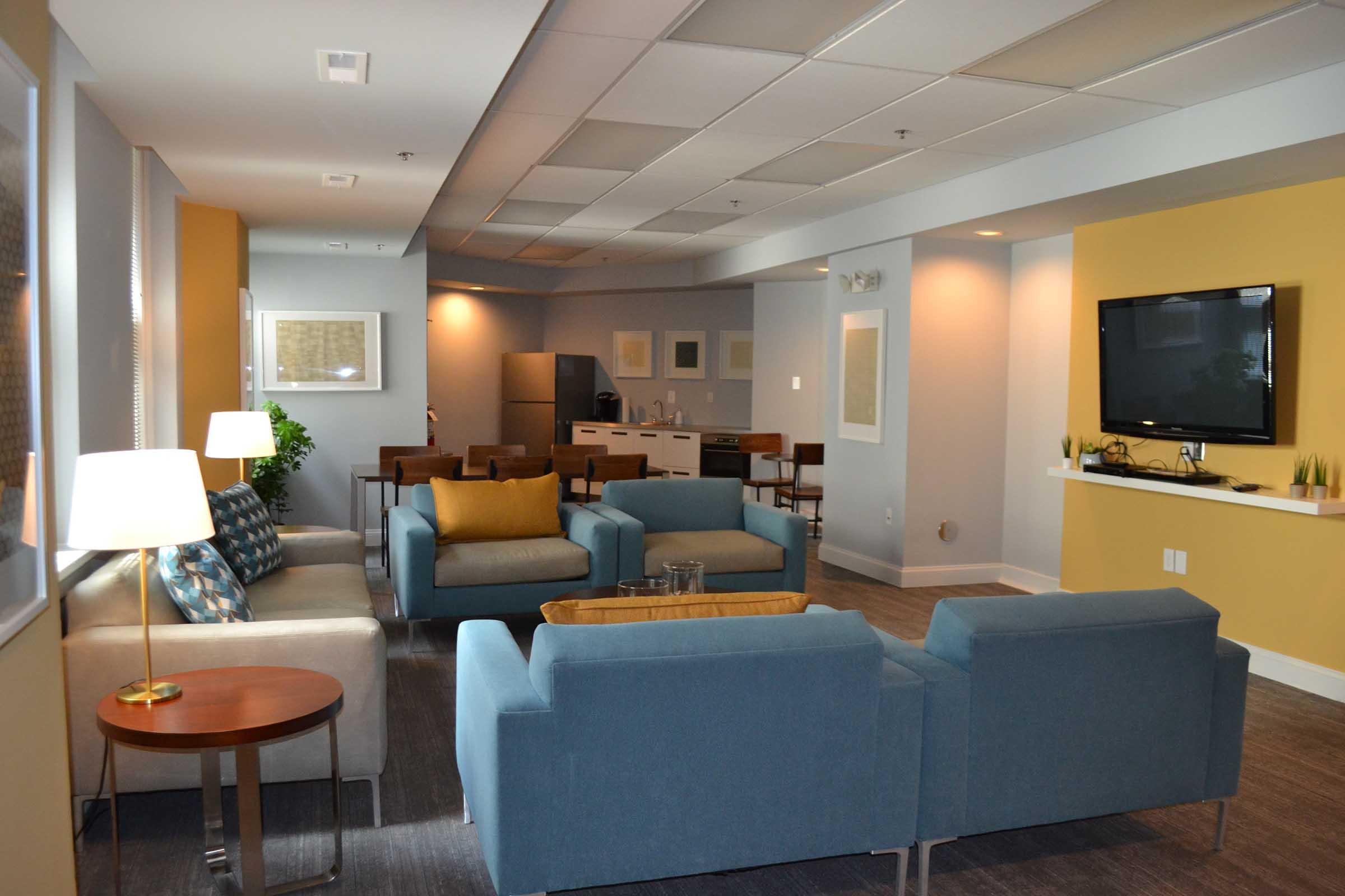 A cozy lounge area featuring modern furniture with blue and beige upholstery, a coffee table, and a floor lamp. The space includes a wall-mounted TV and a kitchenette in the background, creating an inviting atmosphere for relaxation or socializing.