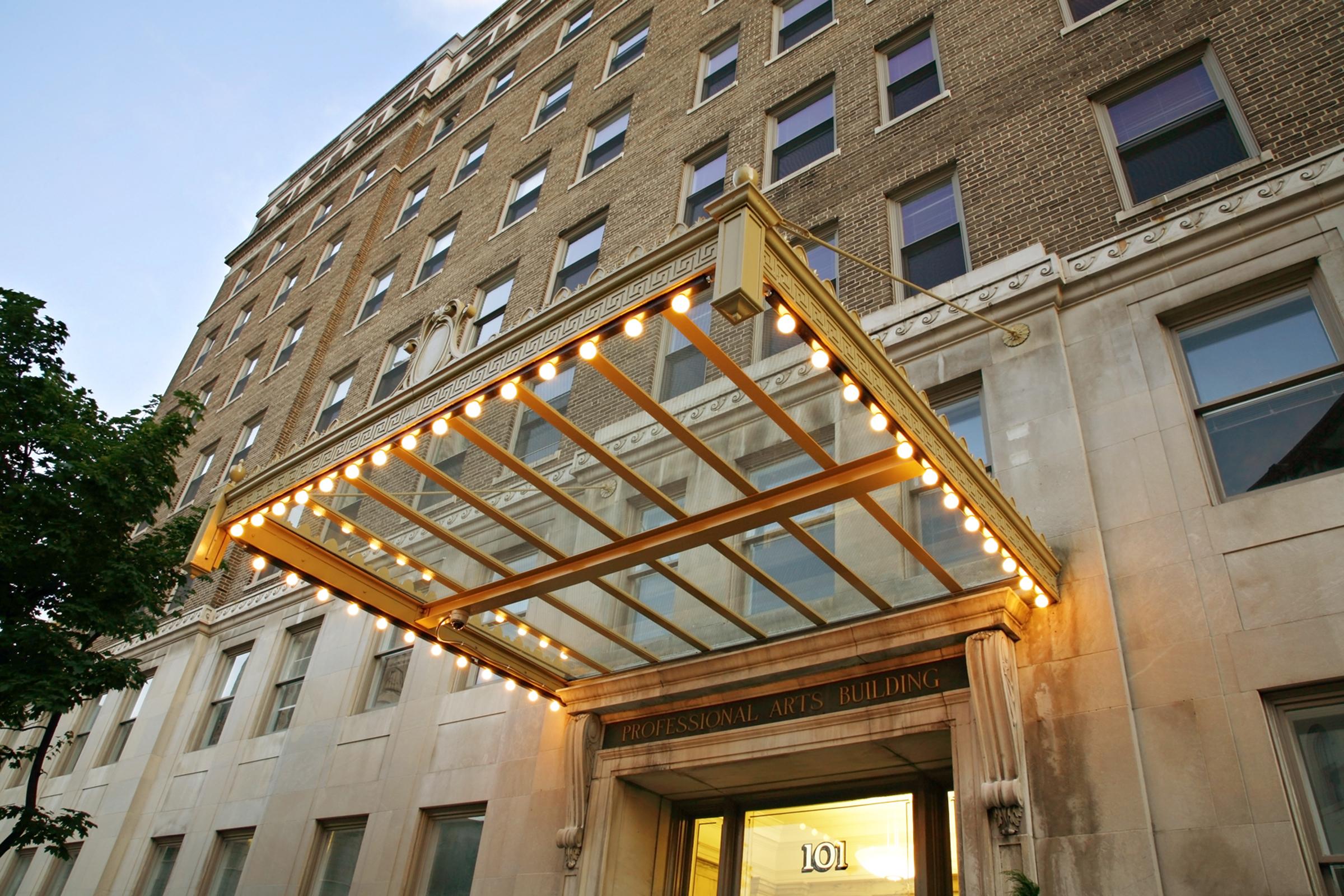 A historic building with a large, illuminated awning featuring string lights over the entrance. The facade is made of beige brick and stone, showcasing classic architectural details. The entrance bears the name "Professional Arts Building" above the door, with large windows visible on the ground floor.