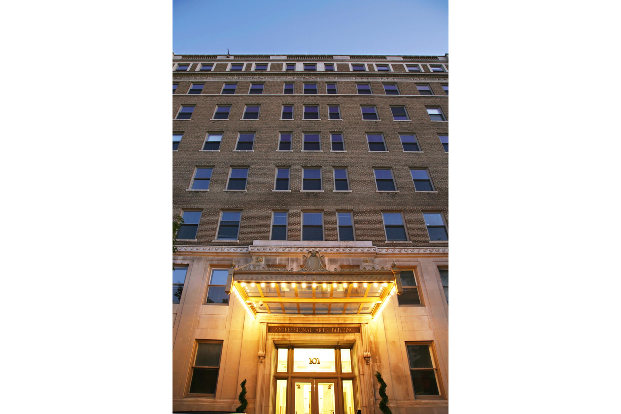 A tall brick building with a large entrance featuring illuminated doors and decorative lighting. The facade includes multiple windows and a classic architectural style, prominently displaying the building number above the entrance. The evening sky is visible in the background.