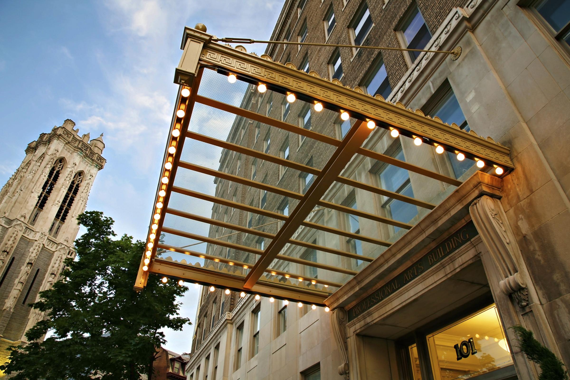 A view of the entrance to the International Arts Building, featuring a modern glass canopy adorned with lights. In the background, a tall clock tower and greenery are visible, set against a historic brick facade. The scene captures a blend of contemporary and traditional architecture.