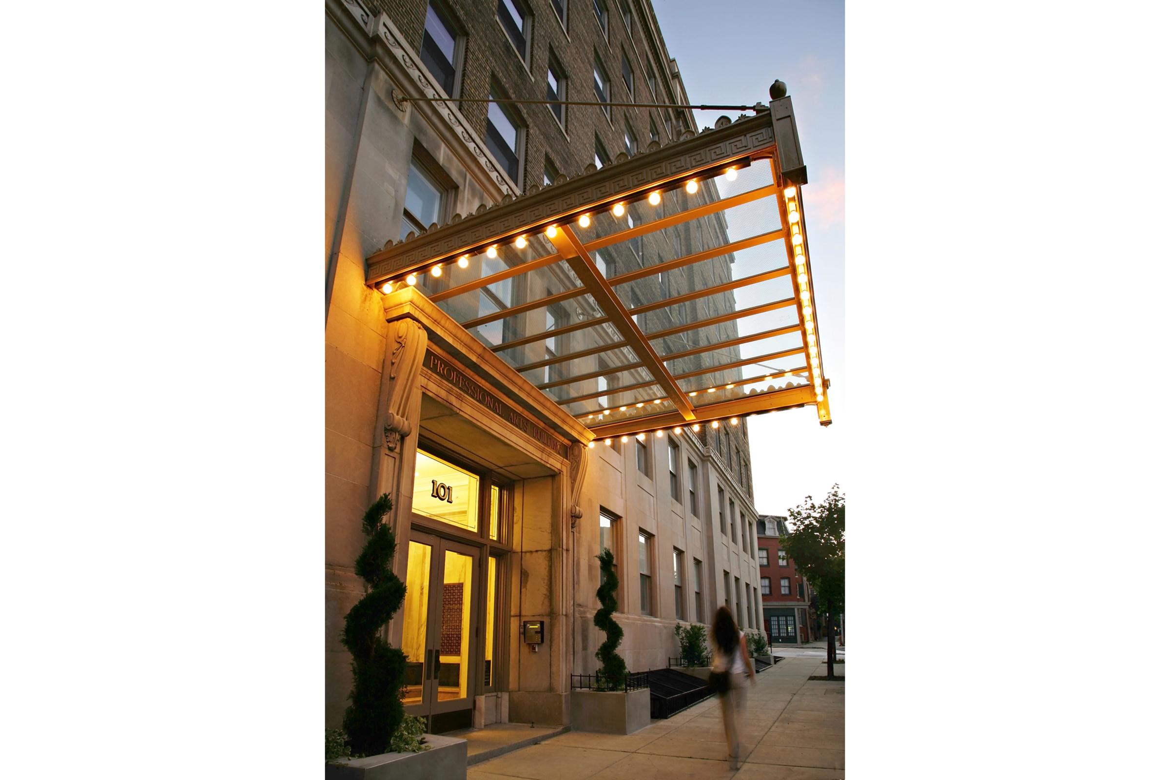 An urban hotel entrance featuring a decorative awning with warm, ambient lights. The entrance is flanked by neatly trimmed plants, and a person walks by on the sidewalk. The building's architecture showcases classic design elements, making it an inviting and stylish entryway.