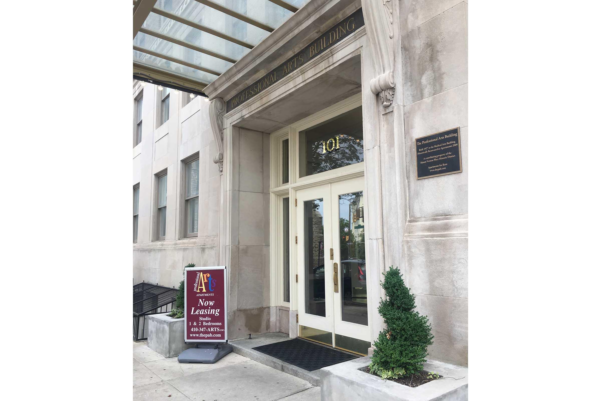 Entrance of the Professional Arts Building, featuring large glass doors with the number 30 displayed. A sign promoting leasing opportunities is positioned on the sidewalk in front, and a small shrub is visible near the entrance. The building has a classic architectural style with an awning overhead.