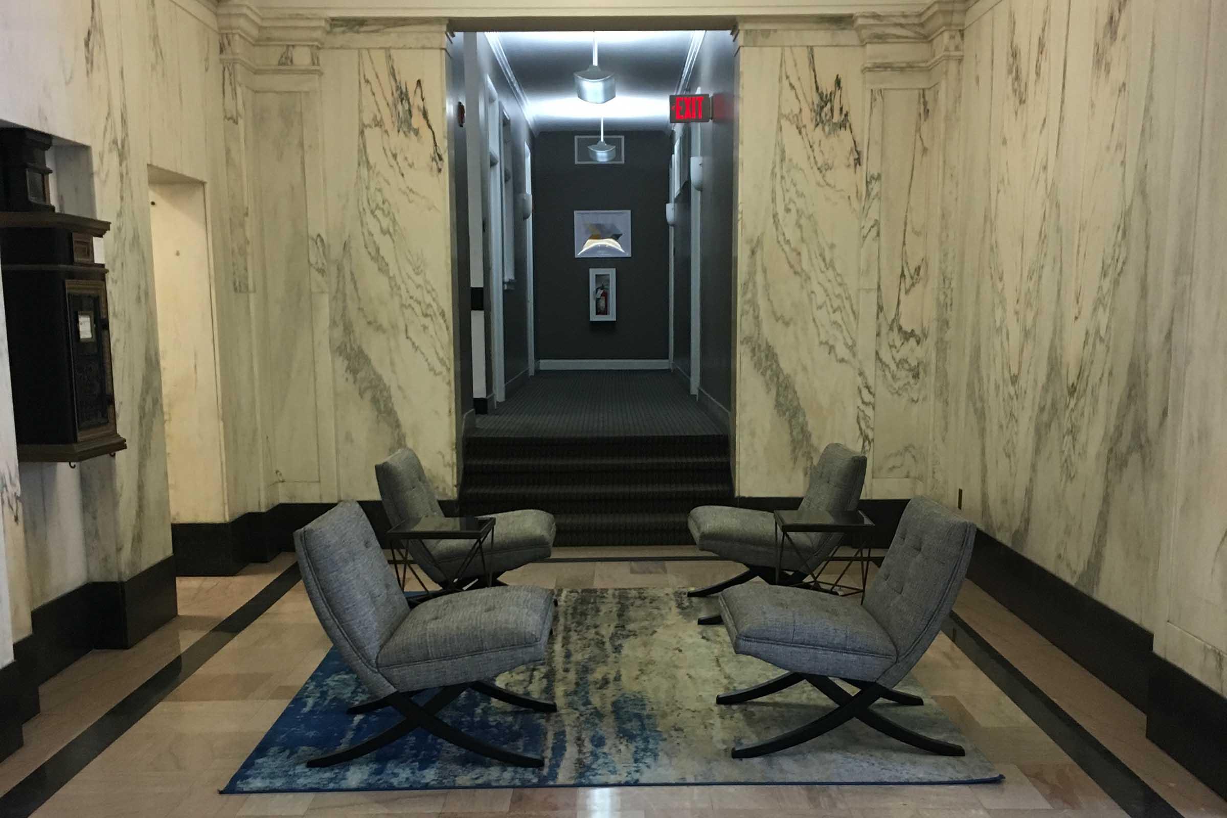 A hallway featuring marble walls and a patterned rug. In the foreground, four grey chairs are arranged in a circle around a small glass table, creating a cozy seating area. The background contains a dark corridor with artwork on the walls and exits at the end. Soft lighting illuminates the space.