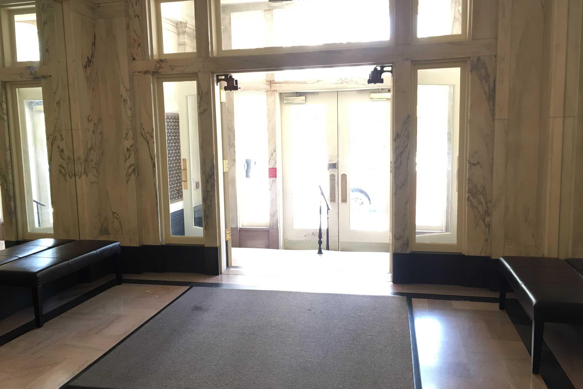 Interior view of a lobby with marble walls, featuring two large glass doors at the entrance. There are benches along the sides and a carpeted area on the floor. Natural light fills the space from the entrance, providing a welcoming atmosphere.