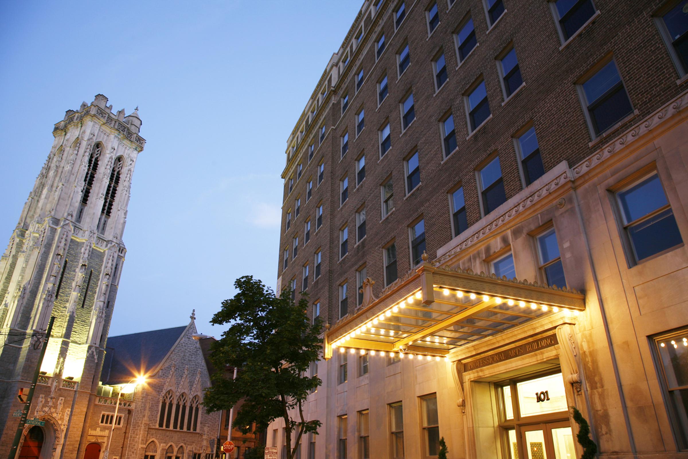 A historic building with elegant architecture, featuring a prominent entrance with lights and a marquee. To the left, a tall bell tower stands alongside a smaller church, surrounded by trees and street lights, under a twilight sky.