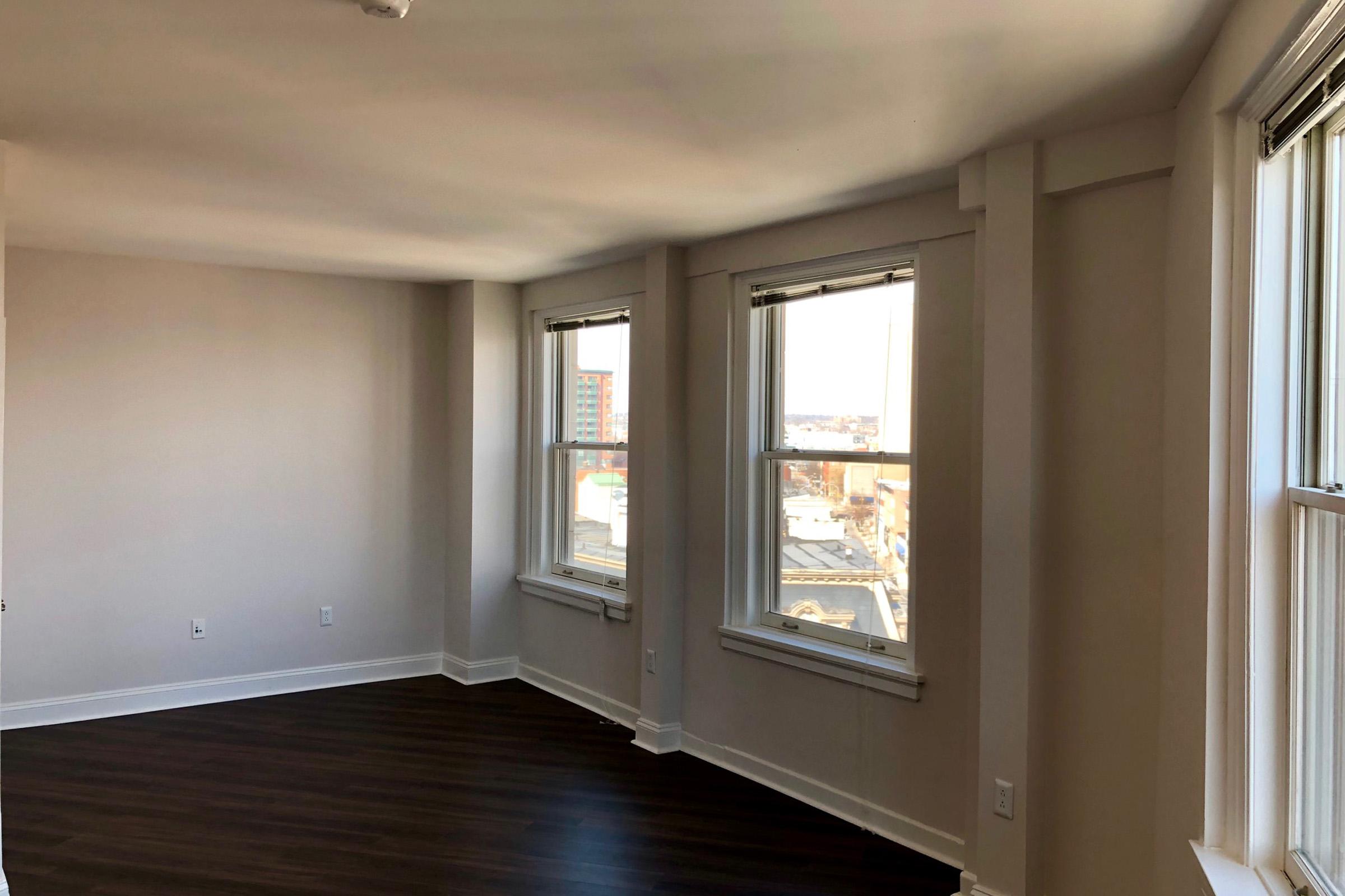 Bright, empty room with freshly painted light gray walls and large windows allowing natural light. The flooring is dark wood, and there's a subtle view of a cityscape outside the windows. The space is uncluttered, suggesting it could be a living room or bedroom in an apartment.