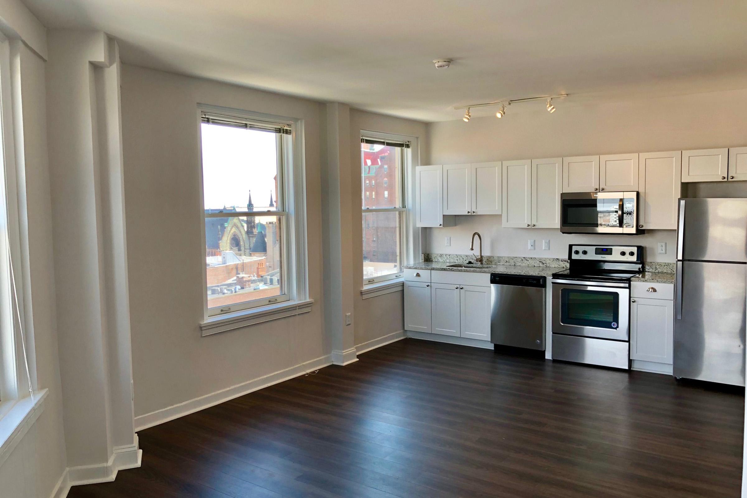 Spacious modern kitchen and living area with white cabinetry, stainless steel appliances, and hardwood floors. Large windows allow natural light in, offering a view of city buildings and skyline. Clean and contemporary design creates an inviting atmosphere.