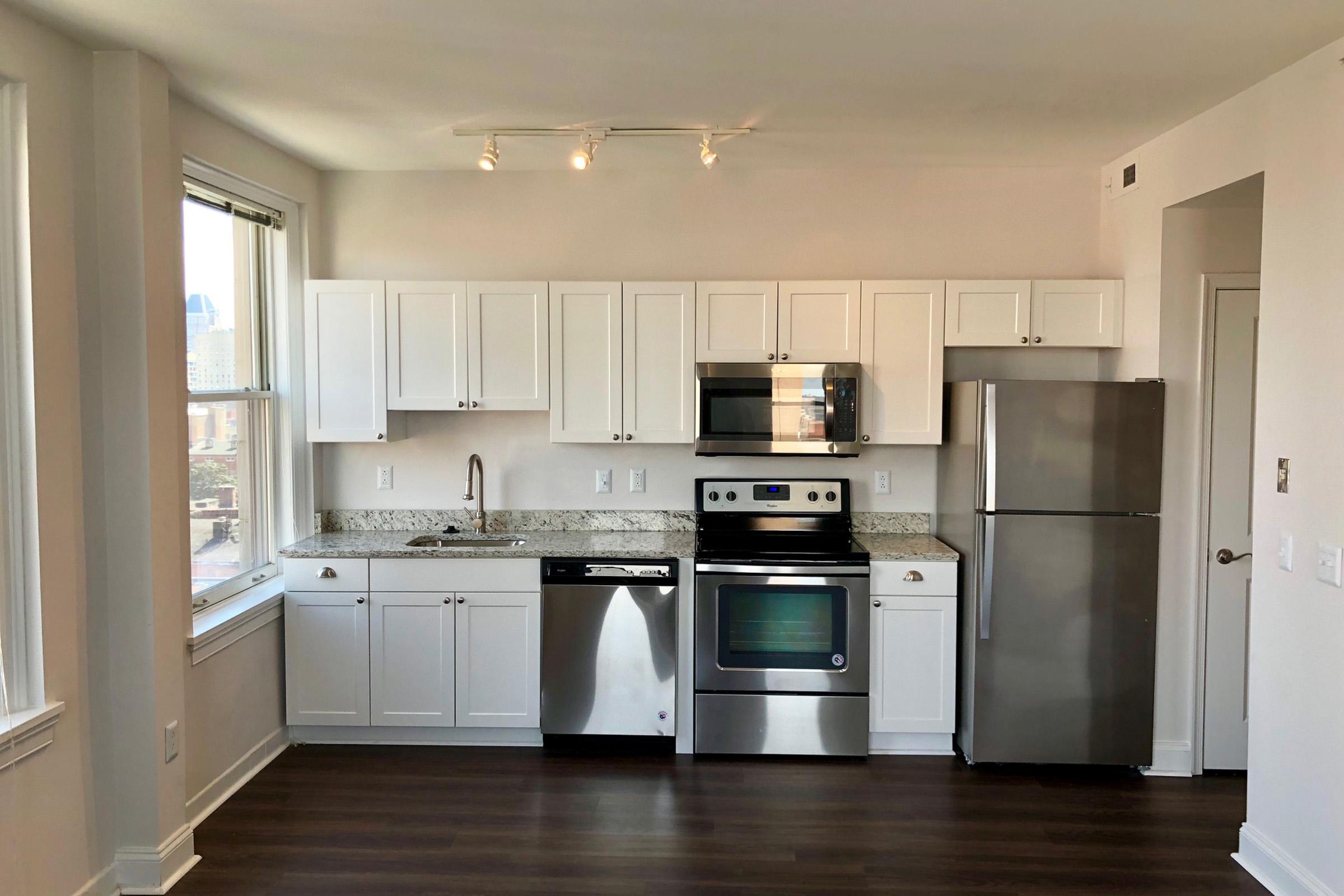 A modern kitchen featuring white cabinetry, stainless steel appliances including a refrigerator, oven, and microwave, and a granite countertop. Large windows provide natural light, with a view of the city visible in the background. The flooring is a dark hardwood.