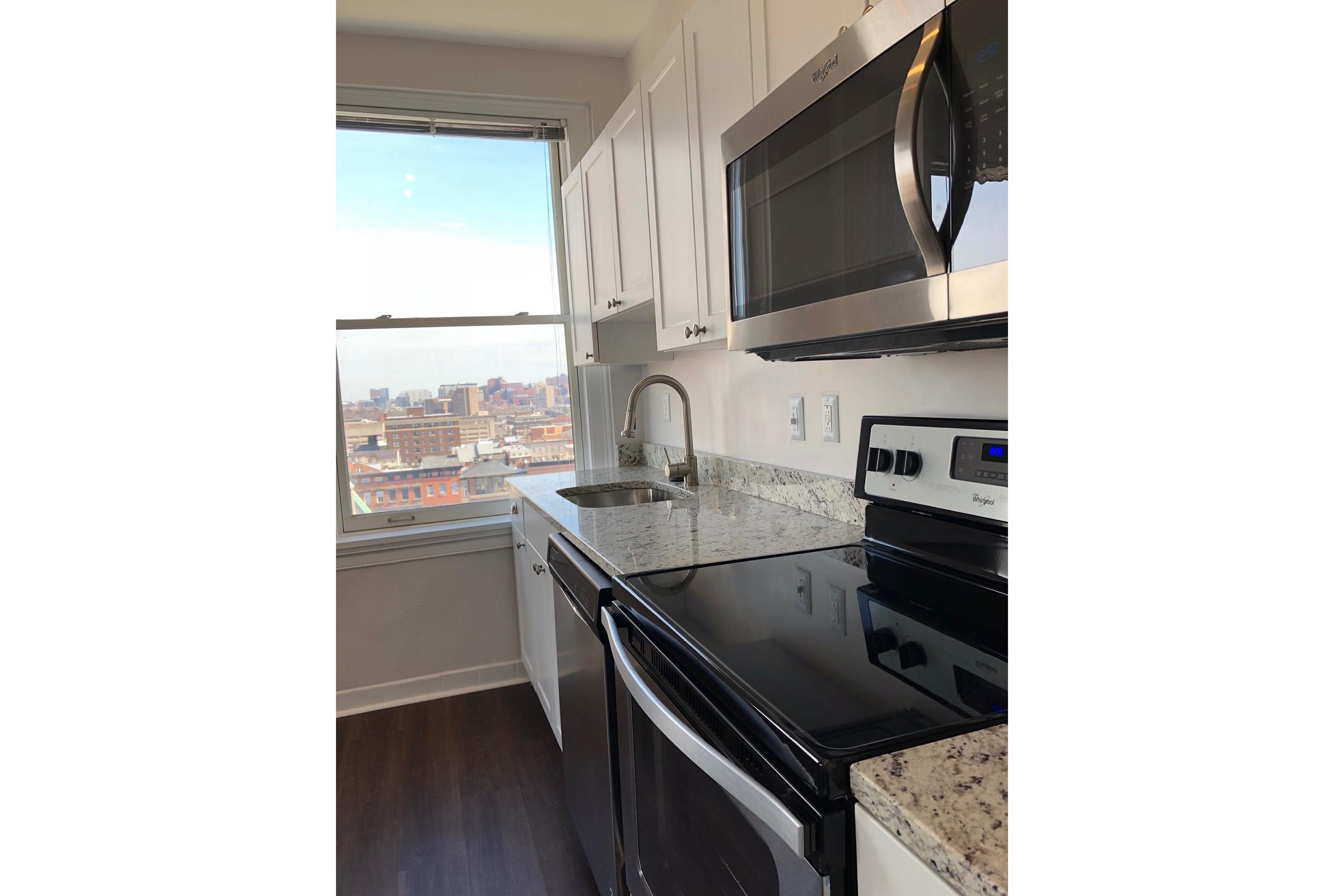 Modern kitchen featuring stainless steel appliances, including a microwave and oven. There is a sink with a sleek faucet, granite countertops, and white cabinetry. A window offers a view of the city skyline. The flooring is dark, enhancing the contemporary look of the space.