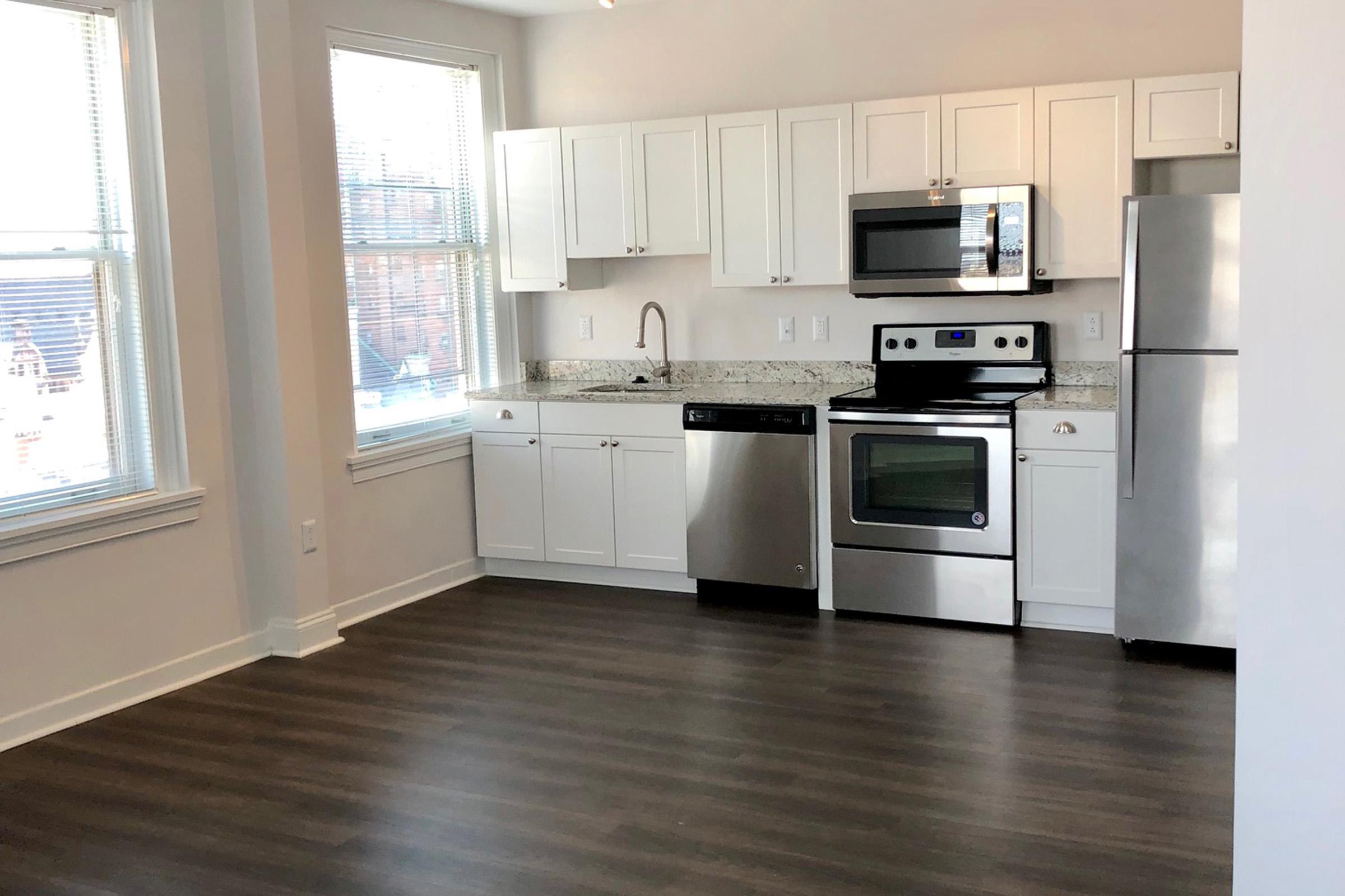 A modern kitchen featuring white cabinets, stainless steel appliances including a microwave, oven, dishwasher, and refrigerator. The countertop is made of speckled granite, and there are large windows allowing natural light to illuminate the space. The floor is covered with hardwood laminate.