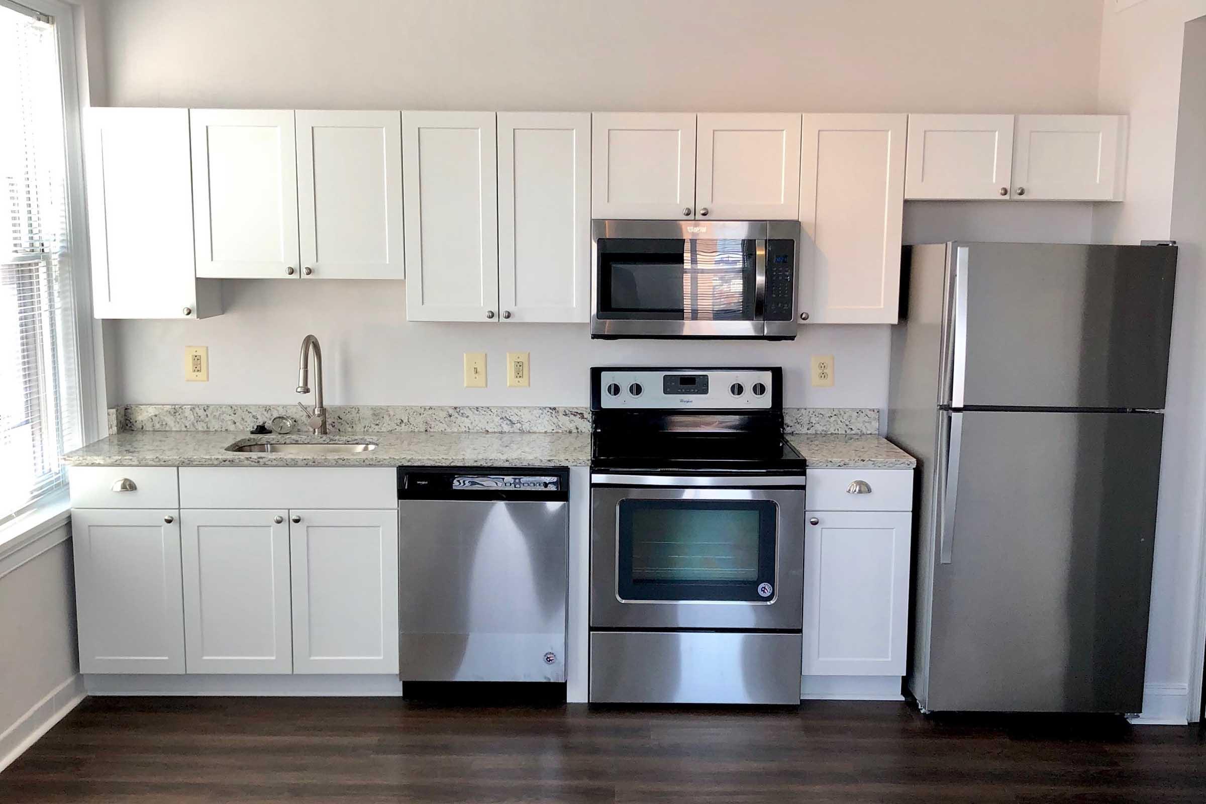Modern kitchen featuring white cabinets with silver handles, a countertop with a light granite finish, stainless steel appliances including a refrigerator, microwave, electric stove, and dishwasher, as well as a stainless steel sink, all set against a backdrop of natural light from a window.