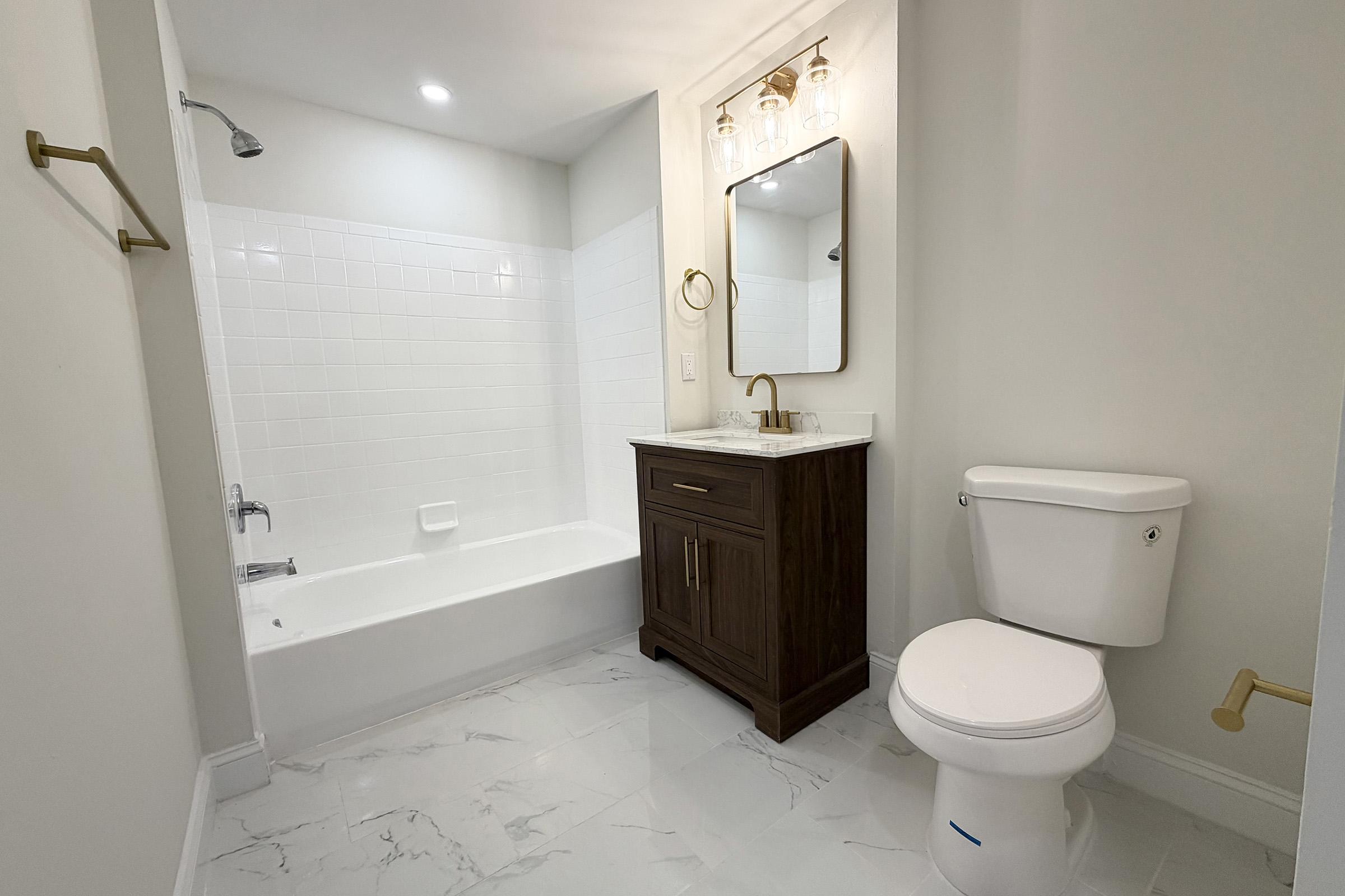 A modern bathroom featuring a white tiled wall with a bathtub, a dark wood vanity with a marble countertop, a round mirror above the vanity, a stylish light fixture, and a white toilet. The floor is made of light-colored tiles, and the space is well-lit with a ceiling light.