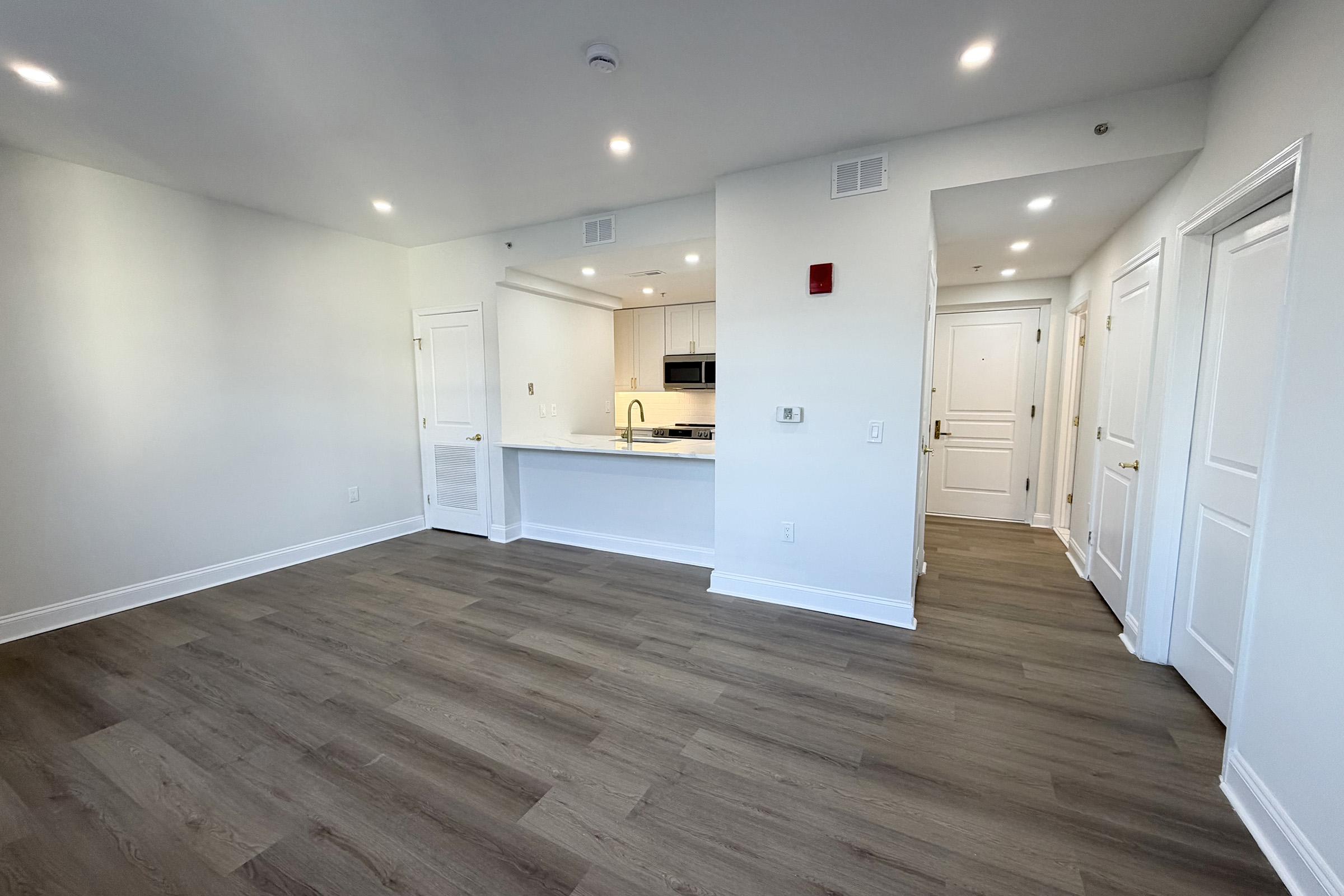 A bright, modern apartment interior featuring light-colored walls and hardwood flooring. The open layout includes a kitchen area with sleek cabinets and appliances. Two doorways lead to other rooms, and there are recessed ceiling lights providing ample illumination.