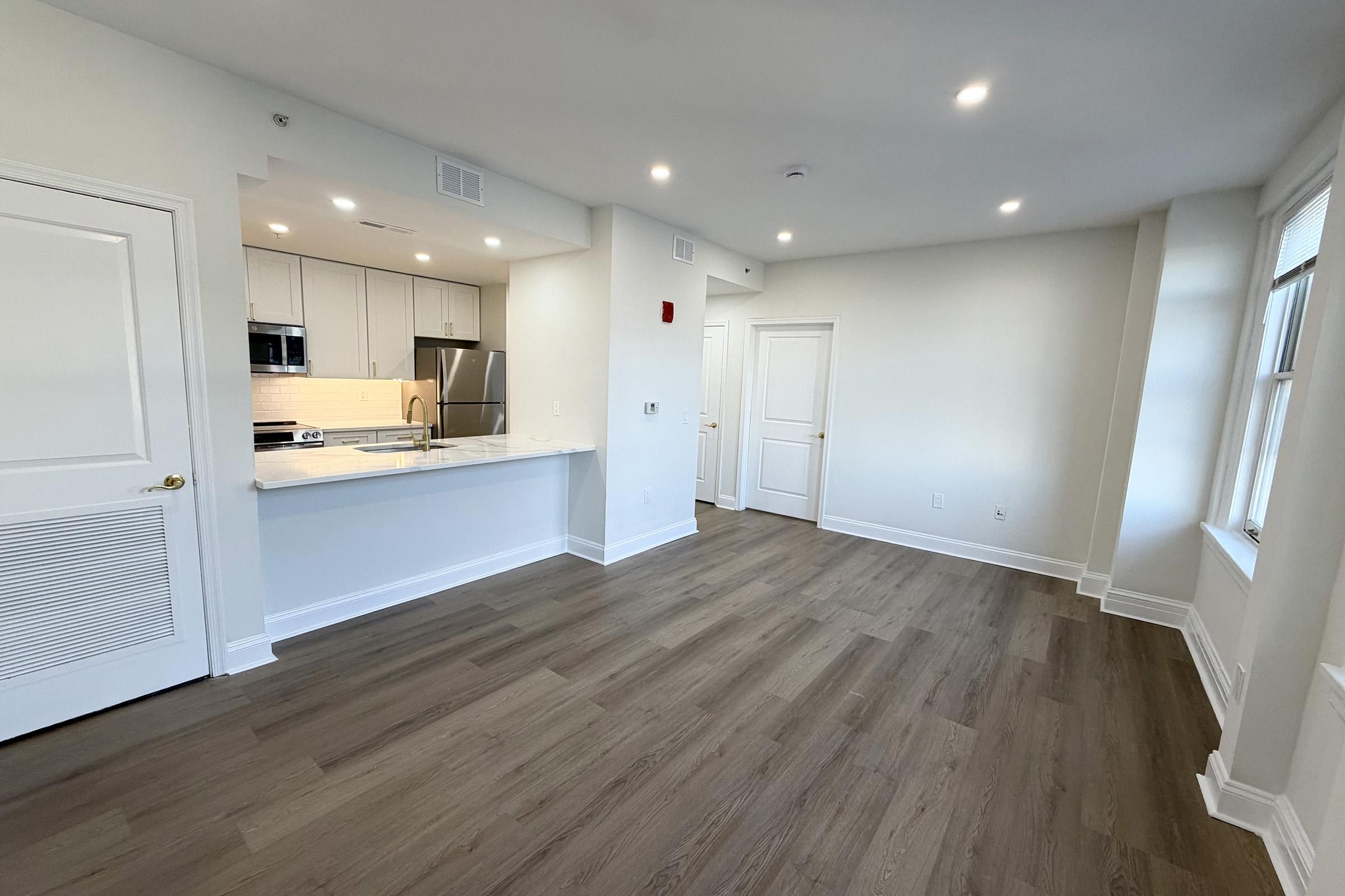 A modern, empty living space featuring a light-colored wall and wooden flooring. The kitchen area is visible on the left with quartz countertops and stainless steel appliances. There are two doors leading to other rooms, and ample natural light comes through the windows. The overall ambiance is bright and spacious.