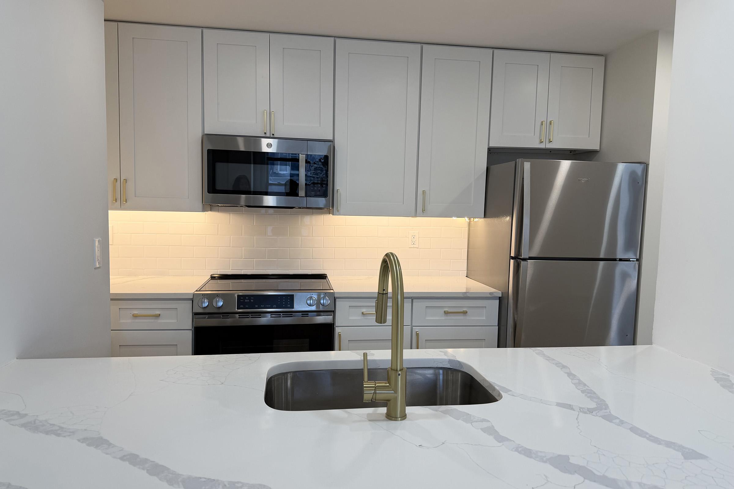 Modern kitchen with light gray cabinetry, a stainless steel refrigerator, and a microwave above a stainless steel oven. A polished countertop features a sink with a brushed gold faucet. Soft lighting enhances the clean and contemporary aesthetic, creating a welcoming cooking space.