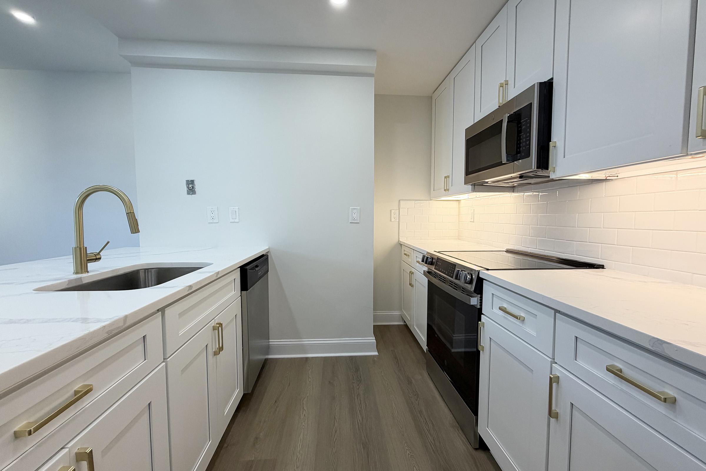 Modern kitchen featuring white cabinets with gold handles, a marble countertop, a stainless steel sink with a gold faucet, and built-in appliances including an oven, microwave, and dishwasher. The walls are a light color, and the flooring is a warm wood tone, creating a bright and inviting atmosphere.