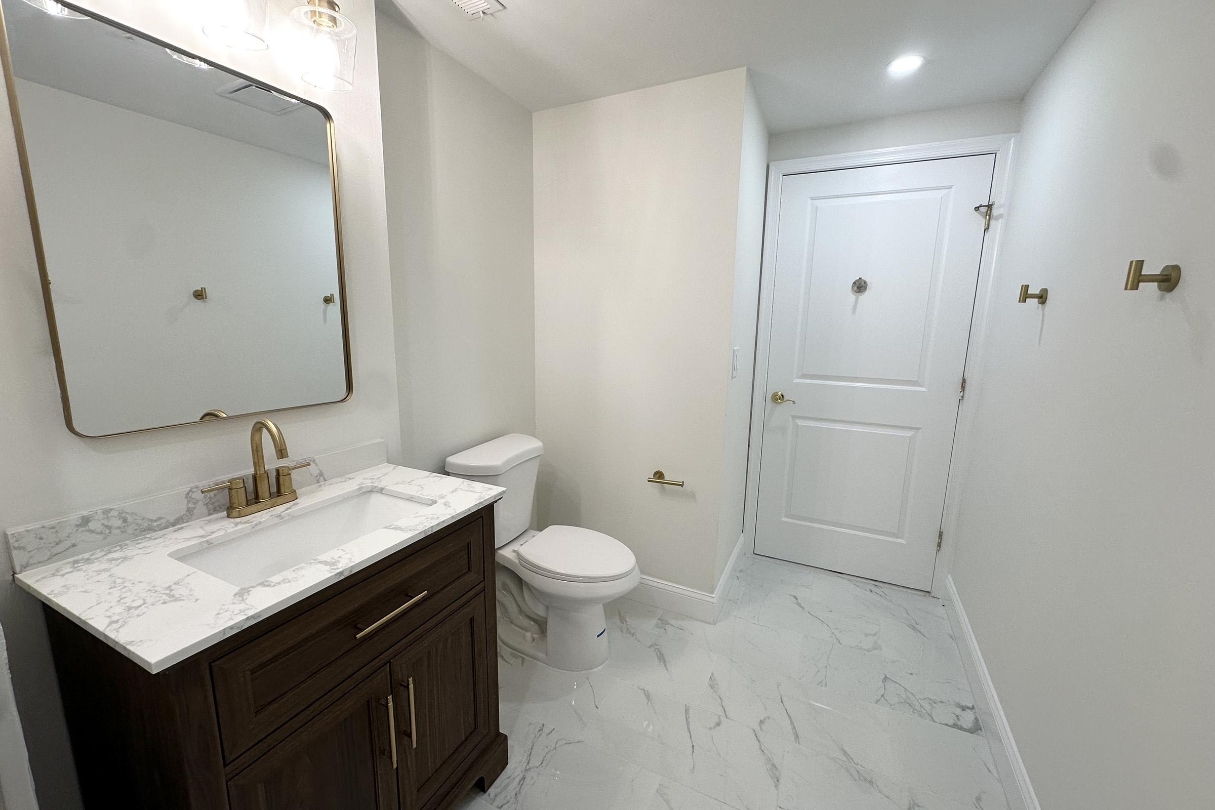 A modern bathroom featuring a white and marble design. The space includes a wooden vanity with a sink and a framed mirror above it, a toilet, and a door leading to another room. The floor is covered in light-colored tiles, and there are gold fixtures and hooks on the walls.