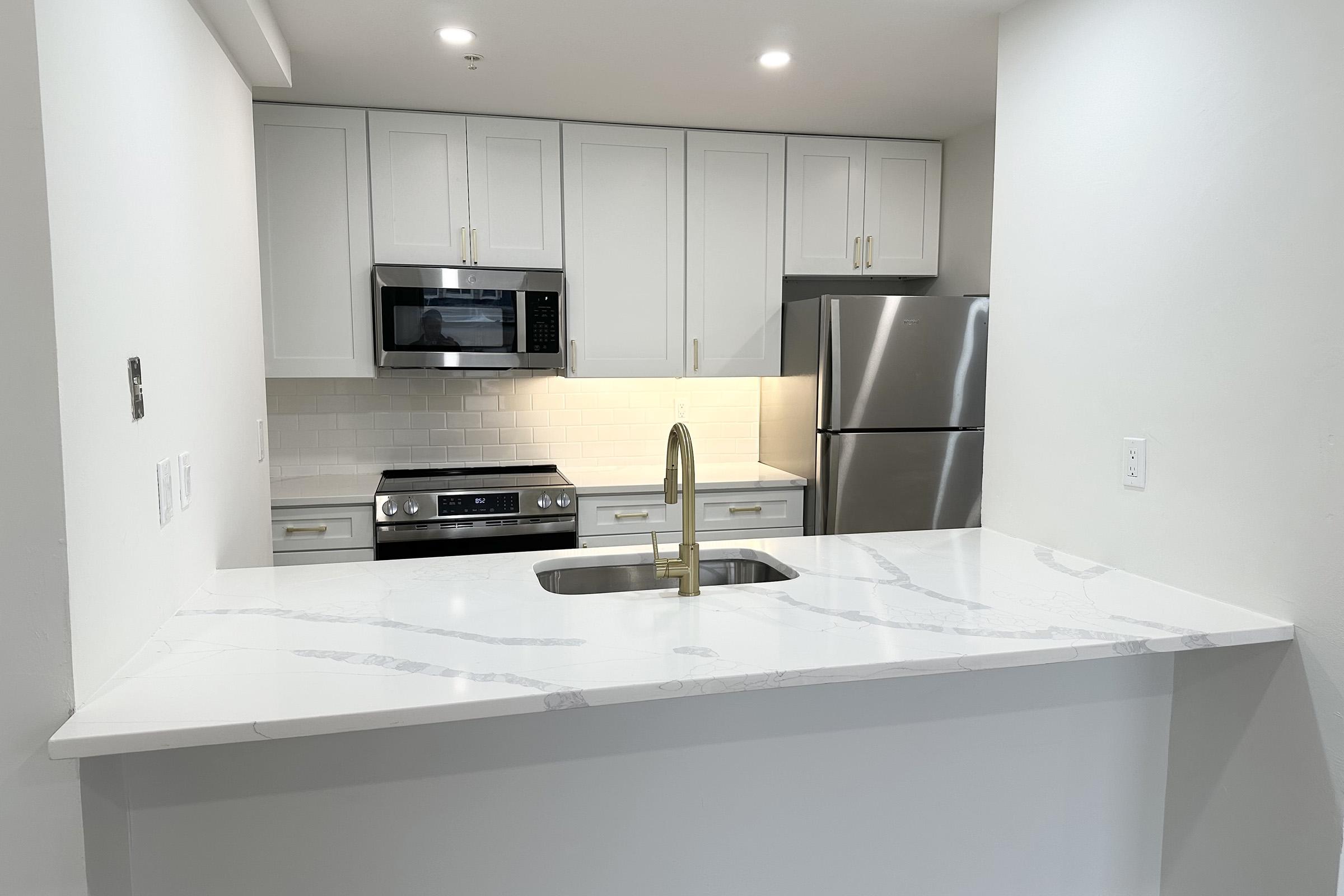 Modern kitchen featuring a white marble countertop with a sink and a stylish gold faucet. The cabinetry is sleek and light-colored, with a microwave and a stainless steel refrigerator visible. Soft lighting highlights the clean design and contemporary appliances.