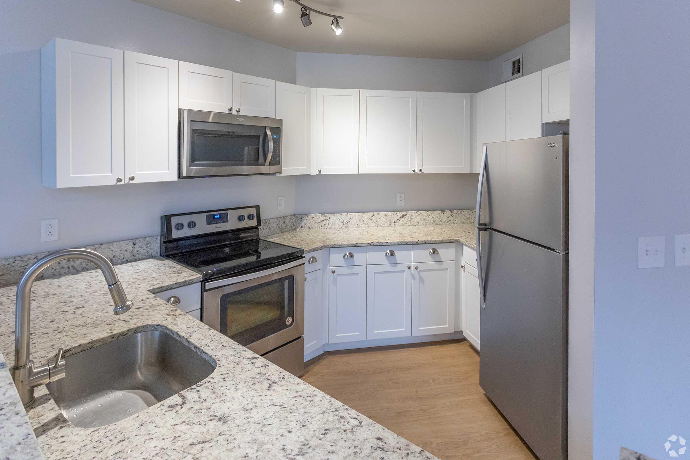 Modern kitchen with white cabinetry, stainless steel appliances including a microwave and refrigerator, and granite countertops. Features a double sink, a stove with an oven, and minimalistic design elements. The flooring is light-colored wood, creating a bright and inviting atmosphere.